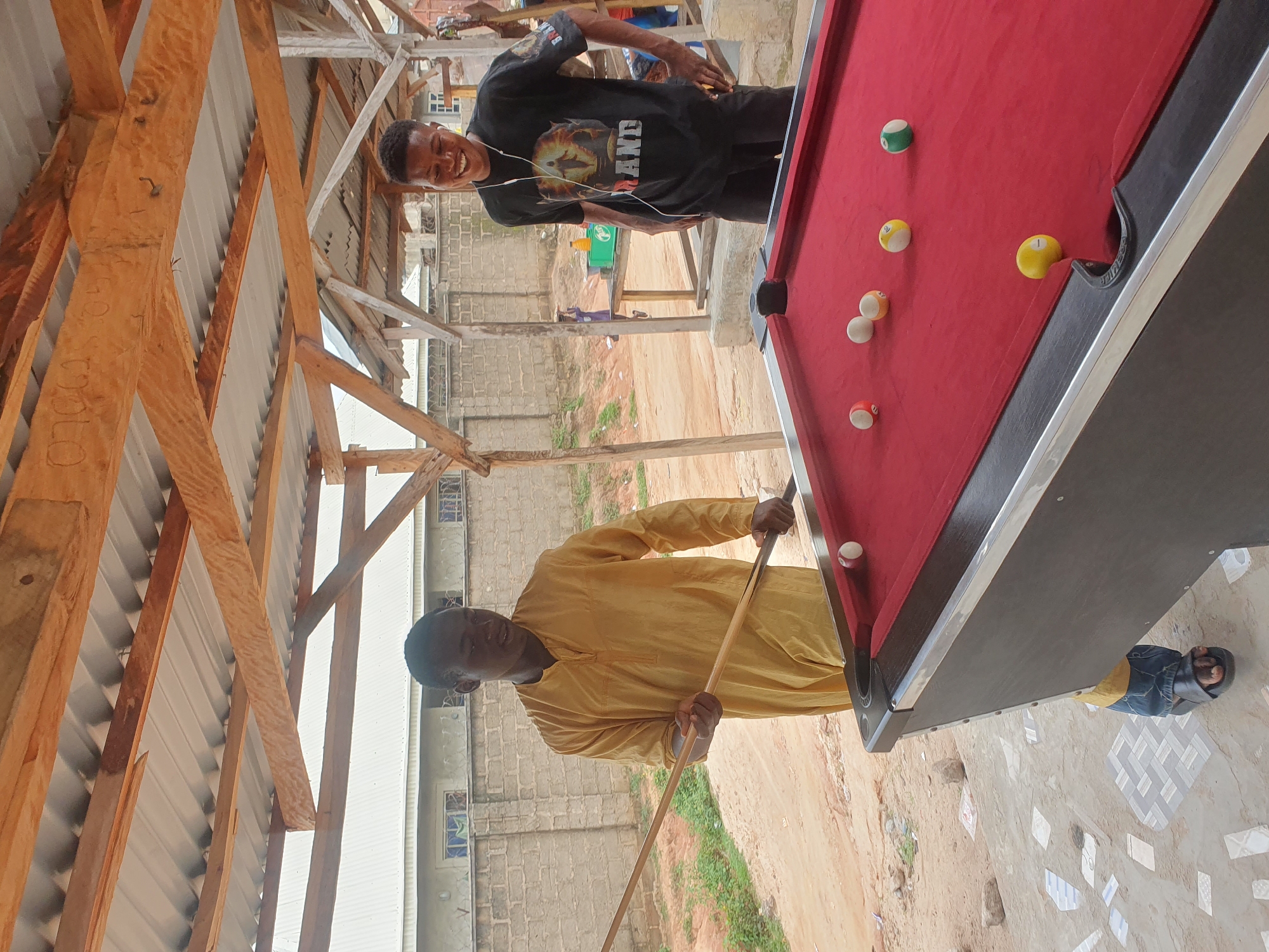 The image depicts an outdoor scene in Lokoja, Nigeria, under a wooden roof structure. Two young men are present. One man, wearing a yellow traditional outfit and blue jeans, stands on the left, holding a pool cue and poised to play. He is positioned next to a red pool table. The second man, on the right, is wearing a black t-shirt with a graphic print and dark pants, and is smiling while listening to earphones. The pool table has several balls on its surface: one white cue ball, a green striped ball, a yellow ball, and two solid yellow balls. The environment is rustic, with a dirt ground and a brick wall visible in the background. The overall impression is a casual recreational setting.