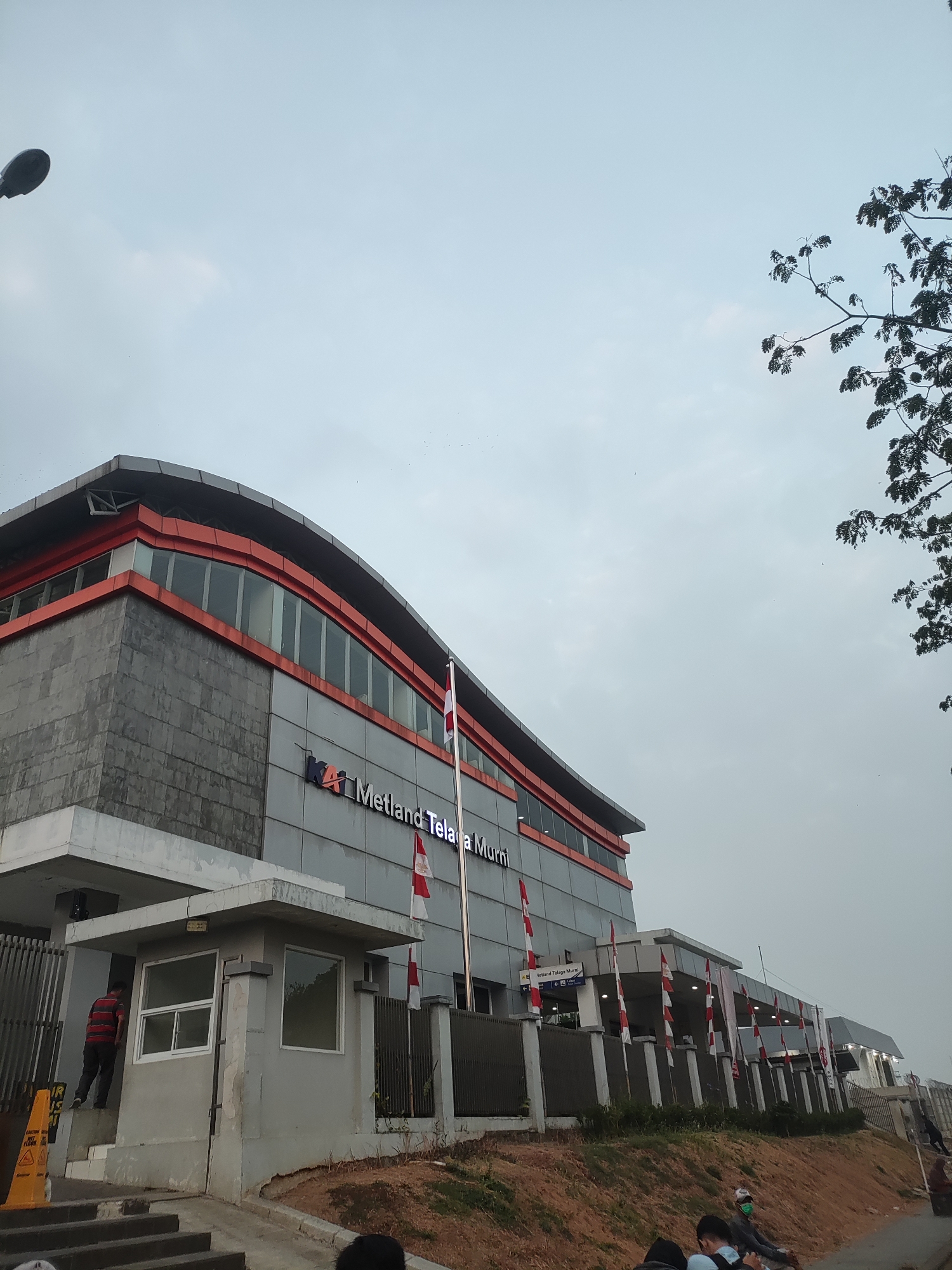 The image depicts the exterior of a modern building complex identified by signage as "Metland Telaga Murni" in Telagamurni, Indonesia. The building features a distinctive architectural design with a grey stone-like facade and prominent orange trim along the roofline and upper window levels. Large windows are visible on the upper floors, reflecting the overcast sky.
In the foreground, a small security post or guardhouse is situated near an entrance. A person wearing a red and black striped shirt is seen standing on steps leading away from the guardhouse. Several Indonesian flags, red and white, are displayed along the building's facade and near entrances, suggesting a national or local celebration or observance.
The surrounding environment appears to be a developed area with paved walkways, a grassy embankment, and fencing. In the background, trees with dark foliage are visible against the pale sky, and a street light is positioned on the left side of the frame. A few individuals are present in the lower portion of the image, some seated and appearing to be waiting. The overall atmosphere is somewhat muted due to the cloudy weather.