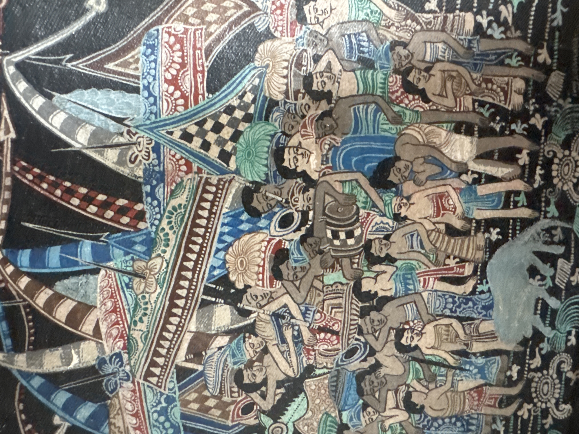 This image depicts a scene with numerous figures rendered in a stylized, folk art manner, possibly a batik painting or textile. The figures, predominantly human, are depicted with simplified features and are adorned in clothing with geometric patterns and bright colors, including blues, reds, and earth tones. They are clustered together in a densely packed composition. Some figures are engaged in activities, such as playing a drum, while others appear to be interacting with each other through gestures and proximity. Overhead, there are abstract shapes that could represent sails or banners, contributing to a sense of movement or procession. In the foreground, a light blue, quadrupedal animal, possibly a sheep or goat, is depicted. The overall impression is one of a communal gathering or celebration. The image does not contain any specific landmarks or details that would allow for its identification as being located in Kuta Selatan, Indonesia.