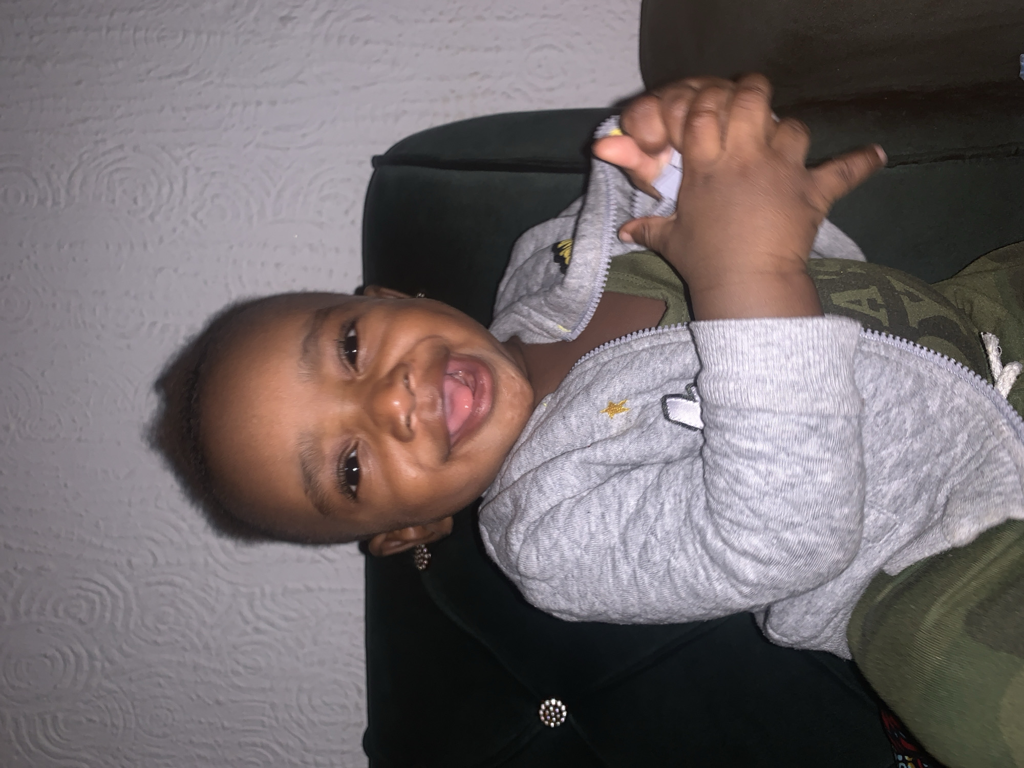 The image shows a close-up of a young Black child sitting on a dark green upholstered chair, indoors. The child is facing the camera, with their mouth open in a wide smile, showing their tongue. They are wearing a grey zip-up hoodie over a green t-shirt, and olive green pants. The background consists of a white textured wall. The lighting is dim, creating a shadow on the left side of the child's face. There are no discernible elements in the image that can be used to determine a specific location within Barakin Akawo, Nigeria.