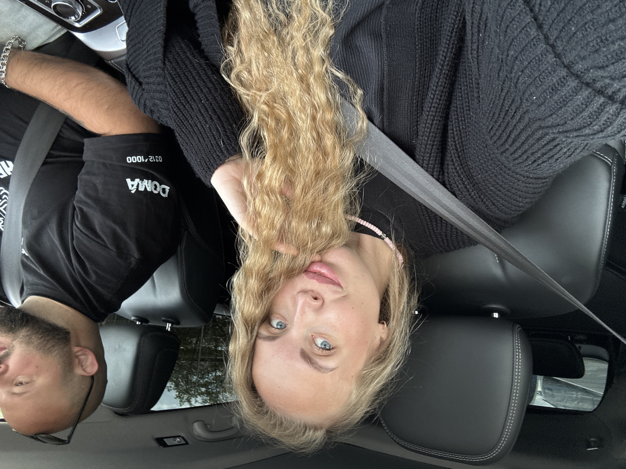 The image shows two people in a car. A man with a beard and glasses is wearing a black t-shirt with the word "DOMA" printed on it. He has a silver bracelet on his wrist. A woman with long blonde hair and blue eyes is also in the car, wearing a black sweater and a pink necklace. Both are wearing seatbelts. The interior of the car is black leather. The location is not discernible as Sochi, Russia, since there are no recognizable landmarks.