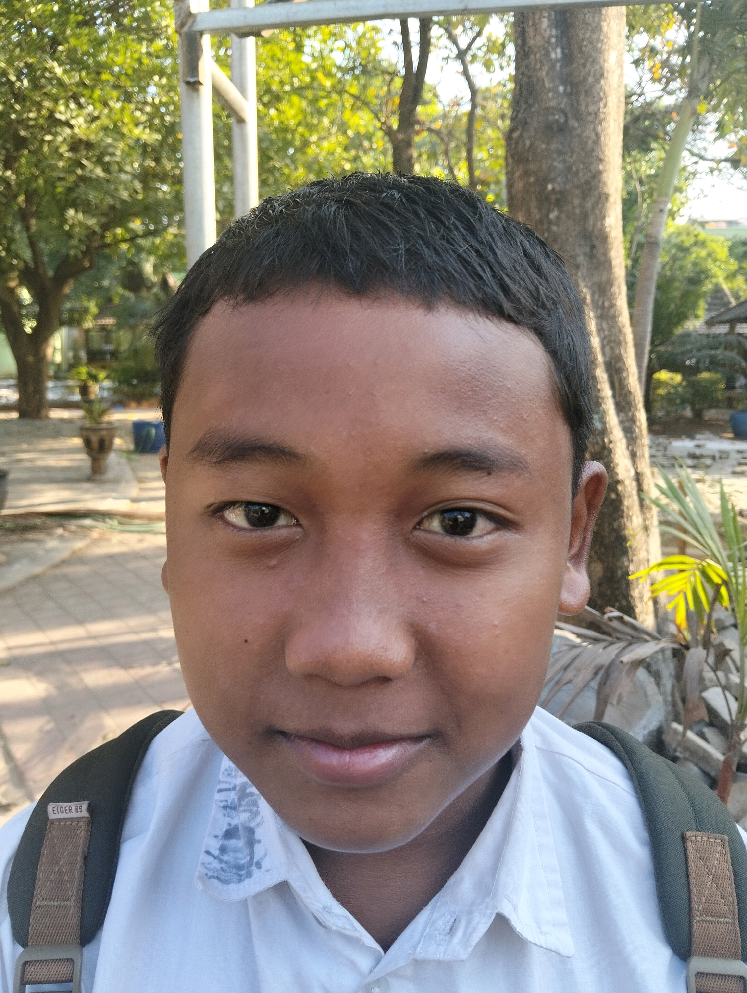 A boy with dark hair, dark eyes, and brown skin is looking directly at the camera. He is wearing a white collared shirt and a dark green backpack strap is visible on his right shoulder. The strap has a tag that says "Eiger 89". The background is blurred, showing trees, a tree trunk, and some foliage. The lighting suggests it is daytime. The image appears to be a selfie taken outdoors. Based on the visible details and common knowledge, this scene could be in Paciran, Indonesia, a coastal district known for its natural beauty and outdoor activities.