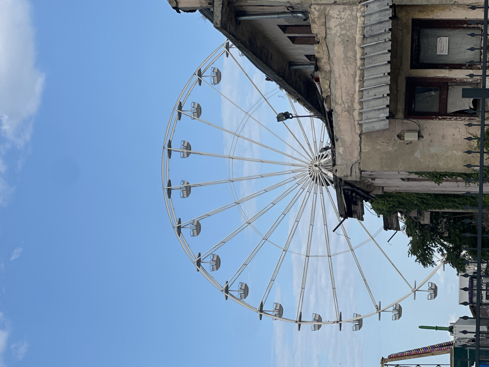 The image shows a Ferris wheel against a blue sky with scattered clouds. In the foreground, there is a dilapidated building with a weathered facade, corrugated metal roofing, and boarded-up windows. The building is partially covered in ivy. A black metal fence runs along the bottom of the image. To the left of the building, a portion of a colorful amusement ride is visible, along with a green structure. The Ferris wheel is white and has numerous gondolas. The scene appears to be an amusement park area that has seen better days, with the contrast between the decaying building and the functional Ferris wheel.