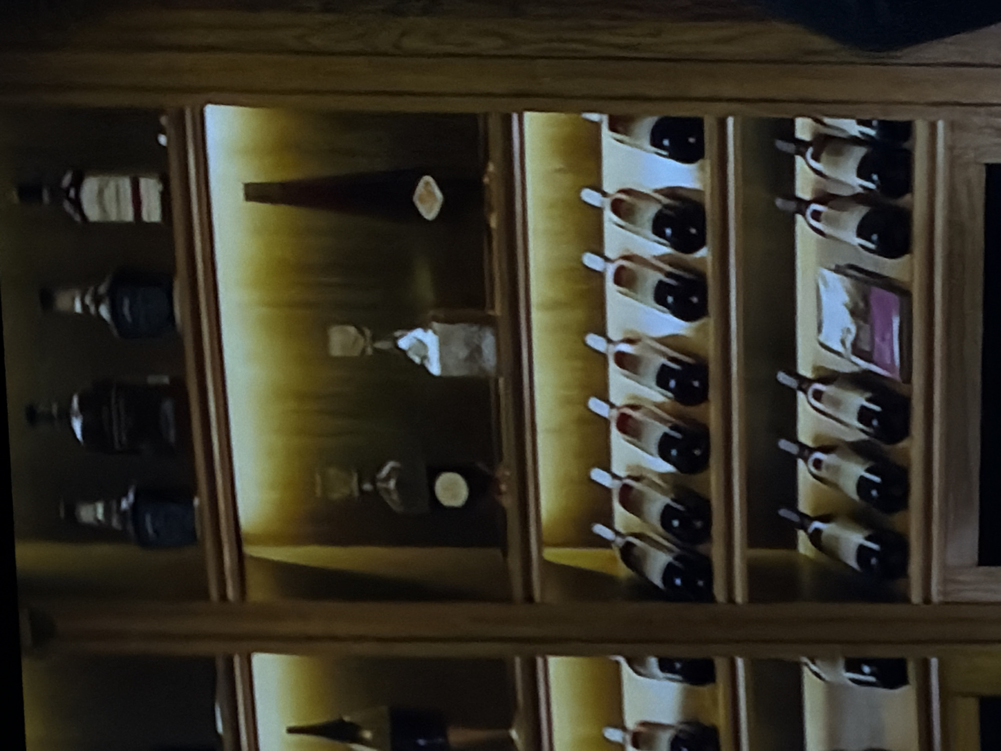 An indoor scene displays a well-organized collection of alcoholic beverages, likely spirits and wine, within custom wooden shelving. The setting appears to be a dedicated storage area, possibly a wine cellar, a bar, or a private collection, located in Sofiivska Borshchahivka, Ukraine.
On the left side of the frame, several tall, upright bottles are visible. The topmost bottle features a distinctive label with a white background and red horizontal stripes. Below it, other bottles with dark liquid and lighter labels are arranged. The central section of the display holds more bottles; one lies horizontally with a light, possibly diamond-shaped detail near its cork, while others stand upright. The right side of the image features numerous wine bottles stored horizontally in individual wooden slots. These bottles are dark-bodied with light-colored labels and caps, arranged in neat rows. One section on the right also contains a small, rectangular object with a purple hue, possibly a box or a special label.
The wooden shelving unit is illuminated by integrated lighting, creating a warm, golden glow on the back panels of the compartments, highlighting the bottles. The overall scene is static, showcasing the collection rather than an activity. No specific text on the labels is legible due to the image resolution. The design of the display suggests careful curation and an aesthetic presentation of the beverages.