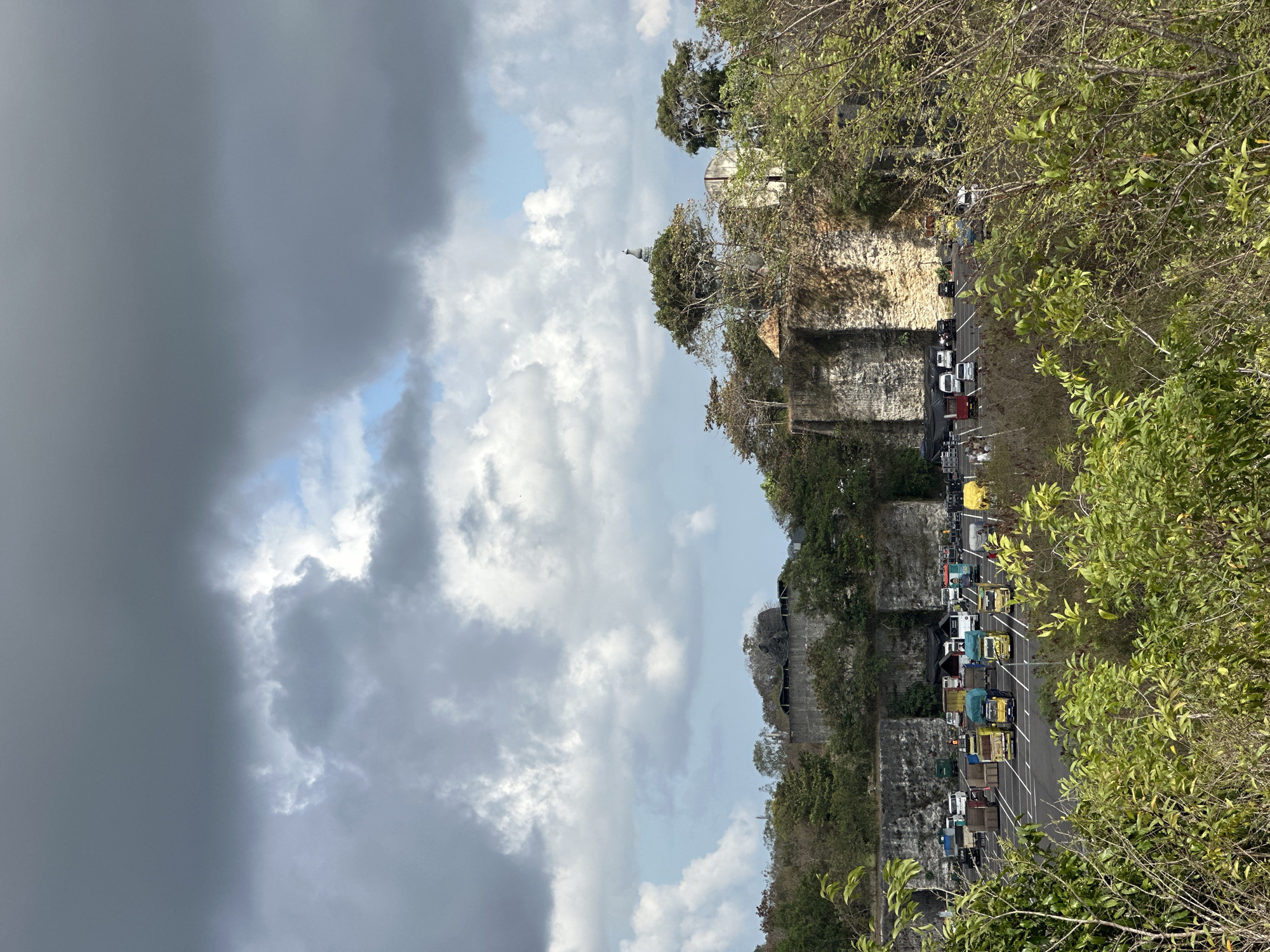 The image, captured in Kuta Selatan, Indonesia, depicts a large, open-air parking lot situated at the base of a cliff face. The lot is filled with a variety of vehicles, predominantly trucks of different colors and sizes, along with several smaller vans and cars. The cliff face behind the parking lot is rough and uneven, with visible rock formations and vegetation growing from its crevices. Above the cliff, a dense canopy of trees and shrubs covers the landscape, and further in the distance, a few structures are partially visible. The sky is partly cloudy, with large, dramatic grey clouds dominating the upper portion of the frame, interspersed with patches of blue sky and brighter white clouds. The foreground is filled with lush green foliage, suggesting a natural, somewhat unkempt environment. The overall impression is one of an industrial or construction-related area integrated into a natural landscape.