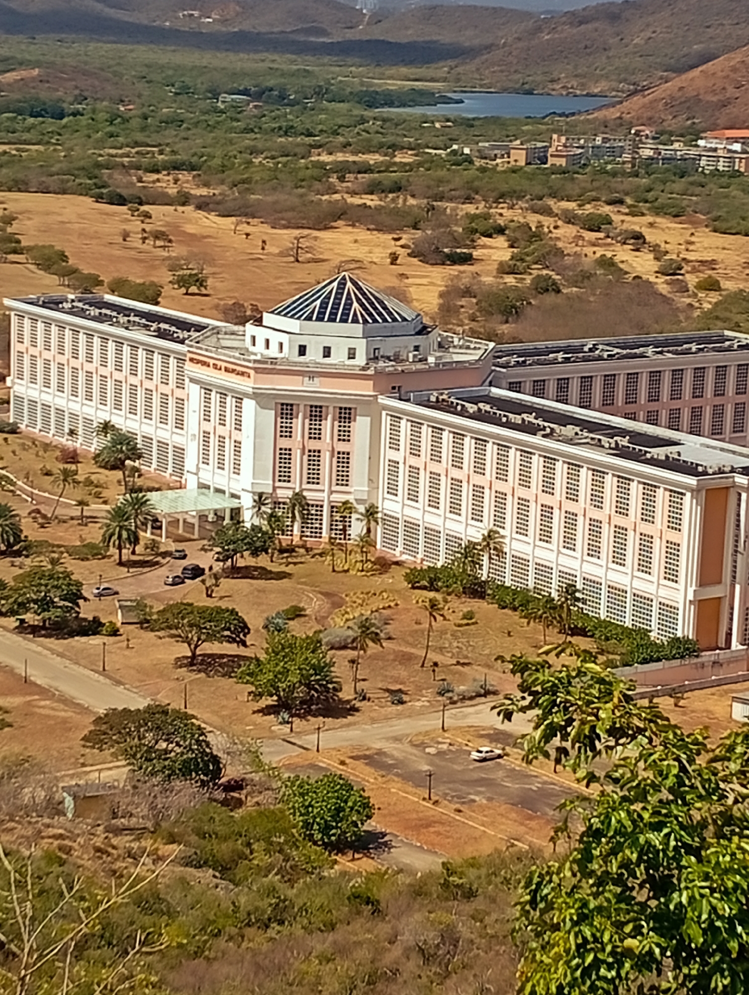 The image presents an elevated view of a large institutional complex identified as Hospital Dr. Rafael Rangel, located within a semi-arid landscape near Juan Griego, Venezuela. The main building is a multi-story structure, primarily white with distinct light pink horizontal banding and pilaster-like elements. It features a grand central section with a prominent, segmented, polygonal dome topped by a clear skylight. Symmetrical wings extend from this central portion, creating a U-shaped layout around a central courtyard. Each wing is characterized by numerous uniformly spaced, grid-patterned rectangular windows.
In front of the central building entrance, a translucent canopy covers a circular drive. Several vehicles are visible: two dark-colored sedans and one lighter-colored sedan are clustered near the entrance, and another white sedan is parked on a paved road further away. The immediate surroundings include landscaped areas with sparse dry, brown grass, green shrubs, and scattered trees, including several palm trees. Paved roads with visible lampposts traverse the property.
The broader environment consists of extensive dry, brown scrubland with scattered green foliage. In the background, a range of rolling hills, partially covered in green vegetation, rises against a clear sky. A body of water, appearing as a lake or lagoon, is visible nestled among the distant hills. Several other smaller buildings with light-colored roofs are discernible in the middle distance. No individuals are clearly identifiable or engaged in specific actions due to the distance and image resolution.
