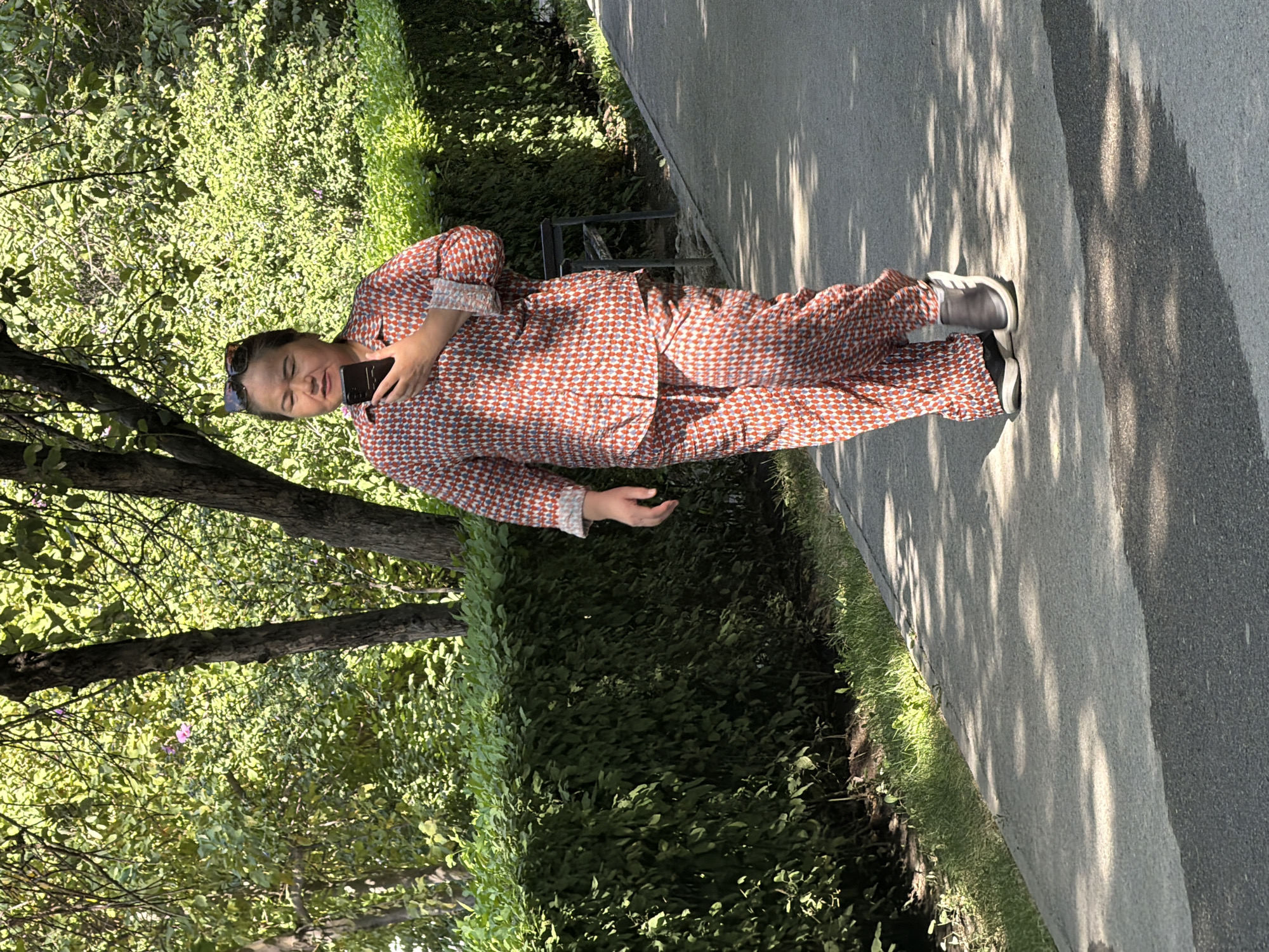 An adult female with dark hair is walking along a paved path in a park or wooded area. She is wearing a red and white patterned pajama set and dark slip-on shoes. She is holding a black smartphone in her right hand and looking at it. Sunglasses are perched on top of her head. The path is bordered by a well-maintained green hedge on the left and trees and foliage on the right. Sunlight filters through the trees, casting dappled shadows on the path. A dark metal bench is partially visible behind the hedge. The overall environment suggests a daytime outdoor scene in a city park.