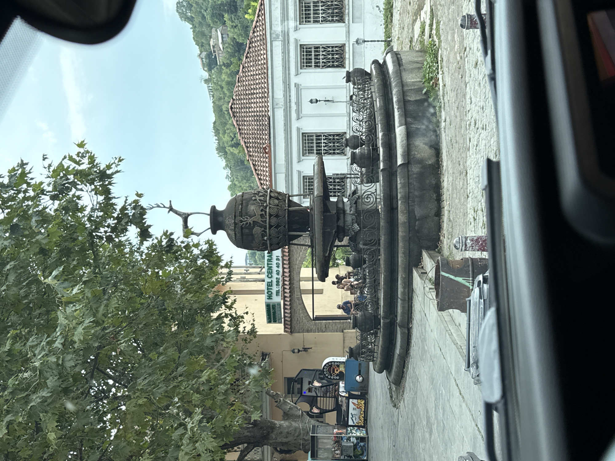 This is a medium-distance, eye-level shot taken from inside a vehicle, looking out at a plaza in Sighnaghi, Georgia. The focal point is a large, ornate stone fountain with multiple tiers and decorative elements, including a metal sculpture of a deer at the very top. The fountain is situated on a cobblestone plaza.
To the left of the fountain, there is a large tree with green leaves providing shade. Behind the tree and to its right, a light yellow building with arched doorways is visible. A sign on this building reads "HOTEL CENTRAL" and displays a phone number. In front of this building, several people are seated at tables, appearing to be patrons.
To the left of the yellow building, partially obscured by the tree, is an outdoor setup that appears to be a stall or small shop selling items, possibly souvenirs or snacks. There are also some amusement-style machines.
To the right of the fountain, a white building with a tiled roof and multiple windows with decorative grills stands prominently. The background shows a hillside covered in greenery with houses scattered on it. The sky is overcast.
In the foreground, the dashboard of the vehicle is visible, including the windshield and part of the steering wheel. Trash receptacles, one a rusty metal bin and another a closed black bin, are present on the edge of the plaza. The overall impression is a public square in a European-style town, likely in Georgia given the context provided.
