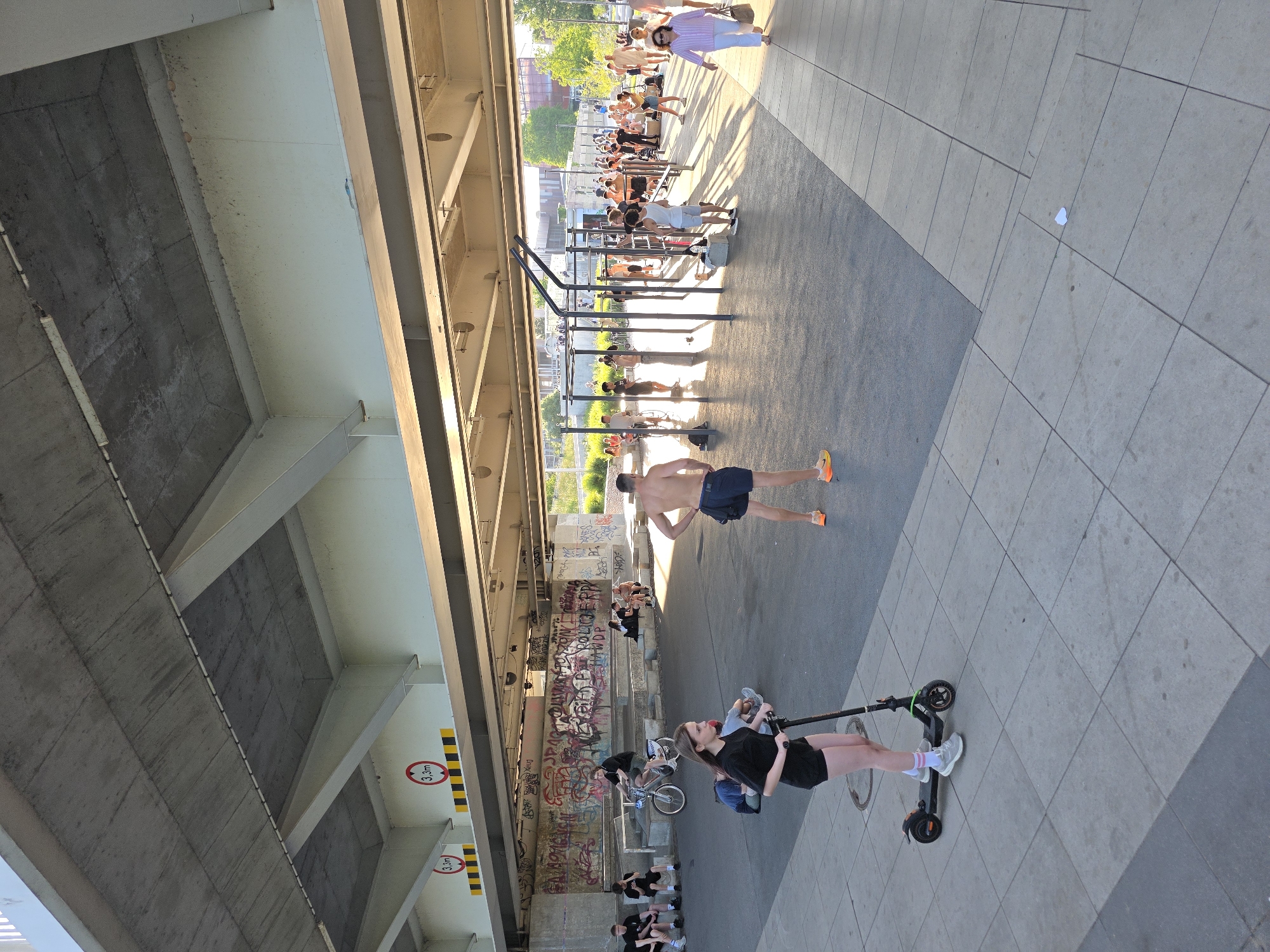 The image depicts an outdoor area beneath a concrete overpass, likely in Warsaw, Poland, given the presence of graffiti in Cyrillic script. The area appears to be a public space with a fitness park containing various exercise equipment, including pull-up bars and parallel bars. Sunlight filters through the overpass, casting long shadows across the scene.
In the foreground, a young woman on a black electric scooter with pink and white socks and grey sneakers is visible. Behind her, a shirtless man with dark shorts and orange sneakers stands facing away from the camera.
Further back, a group of people are gathered around the fitness equipment. Some are exercising, while others appear to be observing or socializing. To the left, a cyclist on a silver bicycle is visible, with a group of people sitting on concrete steps behind him. The concrete steps are covered in graffiti.
In the background, more people are seen walking and standing, and trees and buildings are visible beyond the overpass, suggesting an urban environment. Road signs indicating a height restriction of 3.3 meters are affixed to the underside of the overpass. The ground surface is a mix of concrete pavement and asphalt.