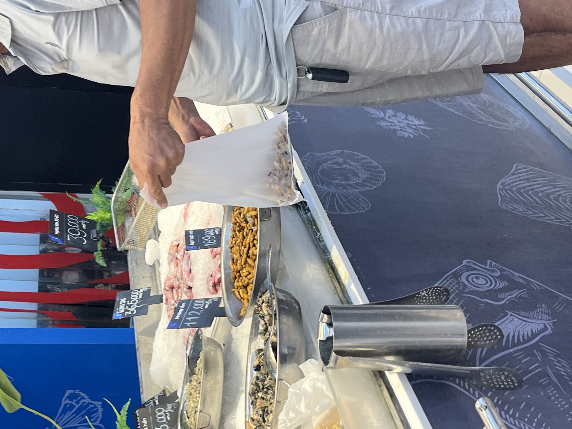 The image depicts a man, wearing a light-colored polo shirt and beige shorts, standing in front of a seafood display at a market. He is holding a white plastic bag containing a food item, possibly dried insects or larvae. The display features various seafood items on ice, with price tags indicating denominations in Vietnamese Dong. One tag reads "365.000" for "Tôm sú". Another tag shows "112.000" for "Nhộng tằm". A third tag displays "189.000" for "Bạch tuộc nhỏ". In the background, red and blue banners are visible. The overall environment appears to be an indoor market with a focus on fresh seafood. The location is identified as Tay Nha Trang Ward, Vietnam.