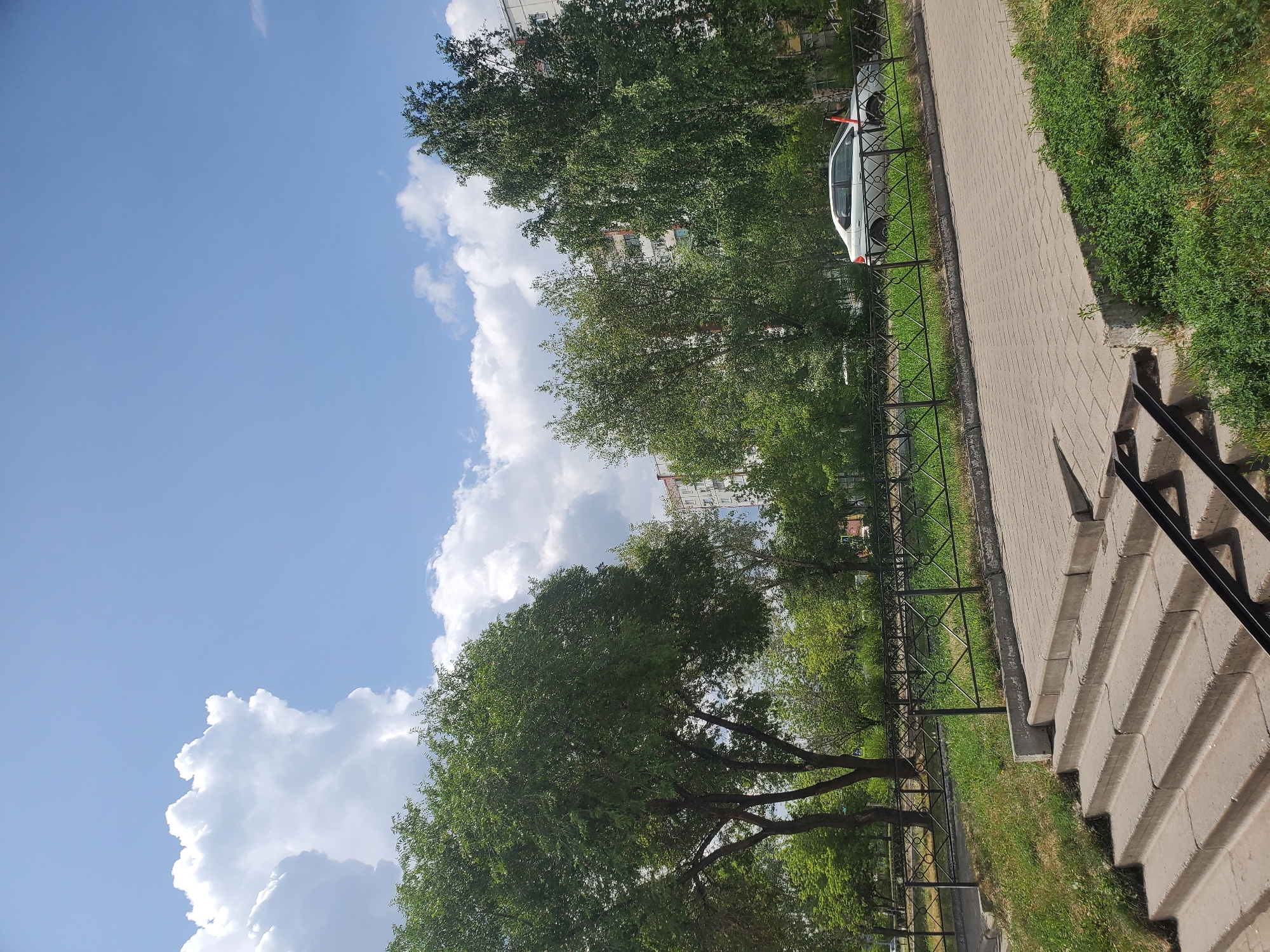 The image captures a bright, sunny day in Sayanogorsk, Russia, with a clear blue sky dotted with fluffy white clouds. In the foreground, a set of concrete stairs leads up to a paved area. A black metal fence runs along a grassy embankment, separating the paved area from a road where a white car is parked. Behind the fence, lush green trees are abundant, with some taller trees providing shade. In the background, multi-story residential buildings are visible, suggesting an urban or suburban setting. The overall scene is one of a pleasant, ordinary day in a residential area of Sayanogorsk.
