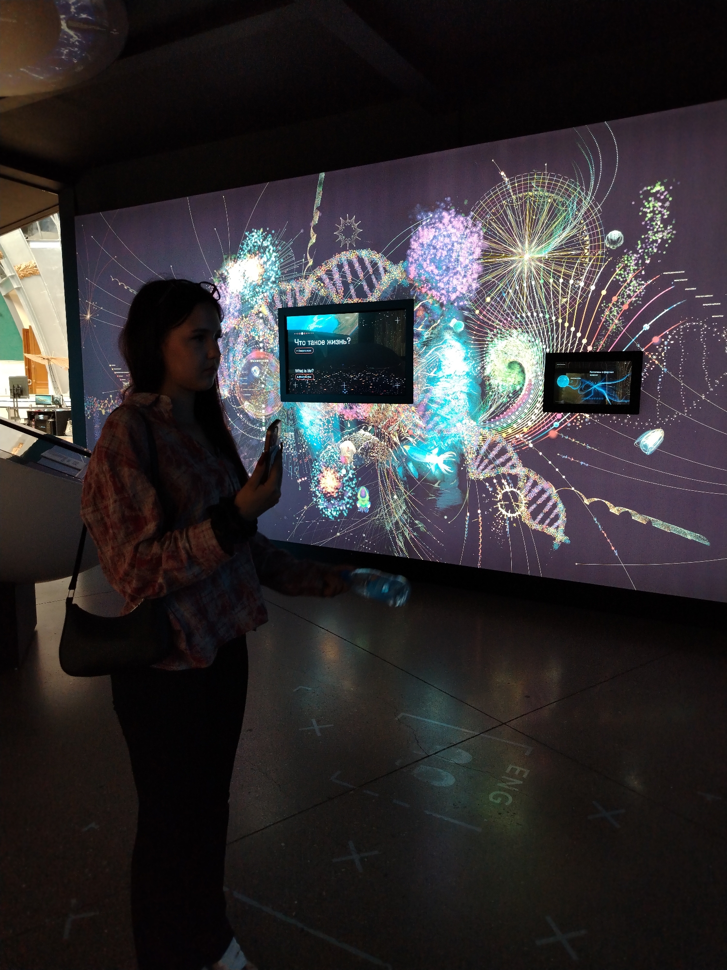 In a dimly lit exhibition space, a young woman stands with her back partially to the viewer, looking at a large, vibrant projection on a wall. The projection displays a complex, abstract visualization of scientific concepts, featuring elements like DNA strands, geometric patterns, and colorful, nebulous shapes. Two smaller screens are embedded within the larger projection. The screen on the left displays text in Russian, which appears to be "Что такое жизнь?" (What is life?) and a button below it. The screen on the right shows a blue graphic of a neuron with radiating lines and text in Russian.
The woman is wearing a plaid shirt and dark pants and carries a black purse over her shoulder. She holds a smartphone in her right hand, its screen illuminated. In her left hand, she holds a clear plastic bottle. The floor of the exhibition space is dark and marked with white lines and cross shapes, possibly indicating exhibit boundaries or interactive zones. Faint projected text that appears to read "ENG" is visible on the floor to the right of the woman. The overall atmosphere is one of curiosity and engagement with the interactive display. The scene suggests an educational or museum setting, likely indoors during the day or evening, given the controlled lighting.