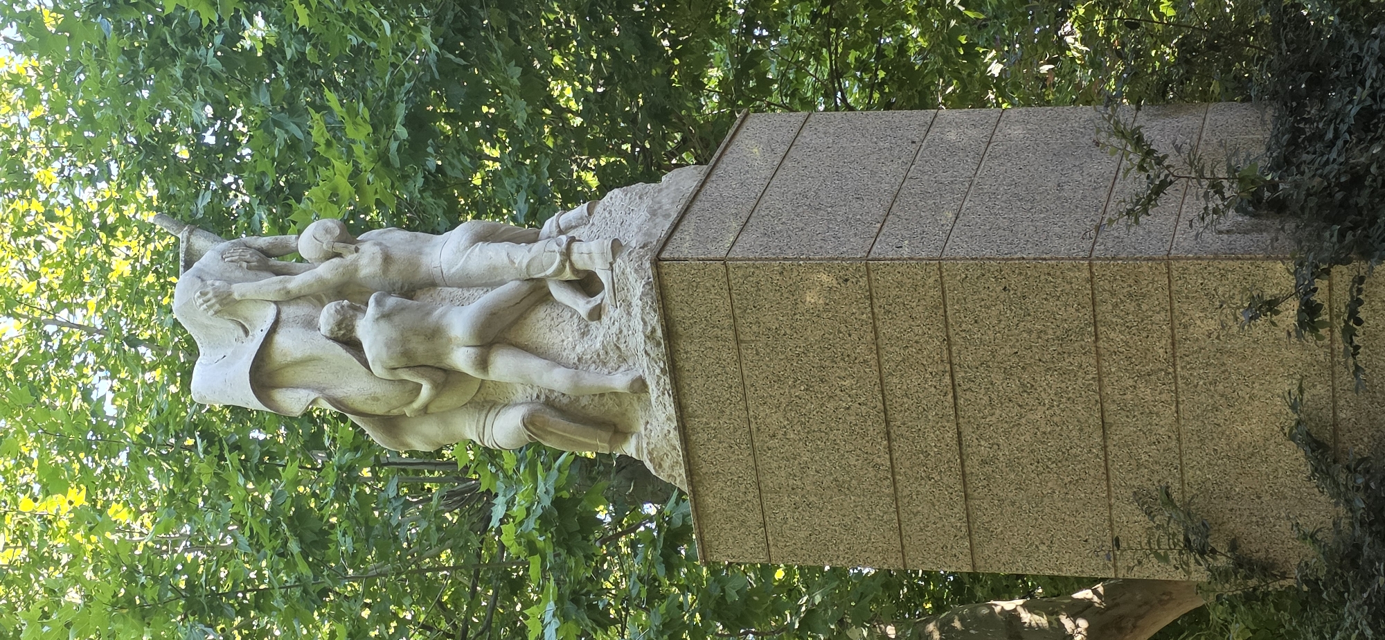 This image depicts a segment of a stone monument featuring a sculptural group of intertwined human figures. The figures appear to be in distress or in a state of struggle, with their bodies contorted and clasped together. One figure, wearing what looks like pants, is lying horizontally, while another above them is leaning over, seemingly supporting or being supported by the others. The sculpture is carved from a light-colored stone with a rough texture.
The sculpture is mounted on a larger, angular base constructed from granite tiles. These tiles are arranged in a pattern, with the top surface angled and the vertical surfaces divided into several rectangular sections by darker grout lines. The base has a speckled appearance, typical of granite.
The monument is situated outdoors, surrounded by lush green foliage. The leaves of the trees are dense, suggesting a sunny day with ample daylight. Sunlight filters through the leaves, creating dappled shadows on the monument and its surroundings. The overall scene evokes a sense of solemnity or perhaps remembrance, given the nature of the sculpture. No visible text is present in the image. The location context provided is Huesca, Spain.