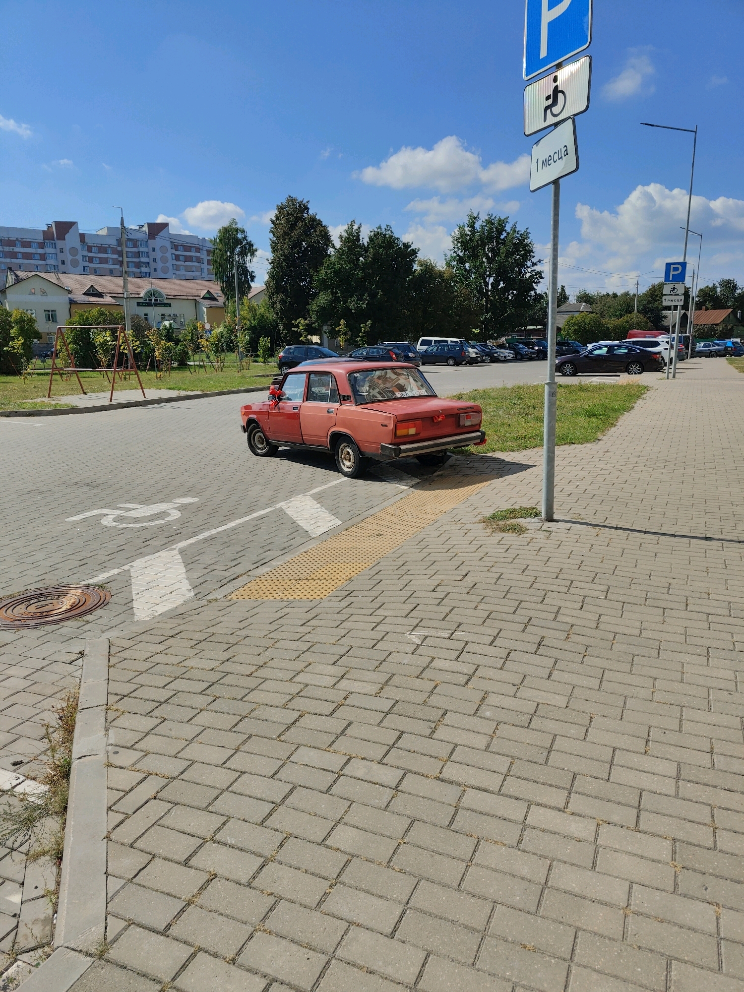 The image shows a red car parked on a cobblestone street in Homyel, Belarus. The car is an older model, possibly a Lada. A sign for disabled parking is visible on a pole nearby, with a second, smaller sign indicating "1 месяц" (1 month). In the background, there are buildings, trees, and other cars parked. The sky is blue with some clouds. There are no people visible in the image. The scene appears to be a typical street view in a residential or commercial area.