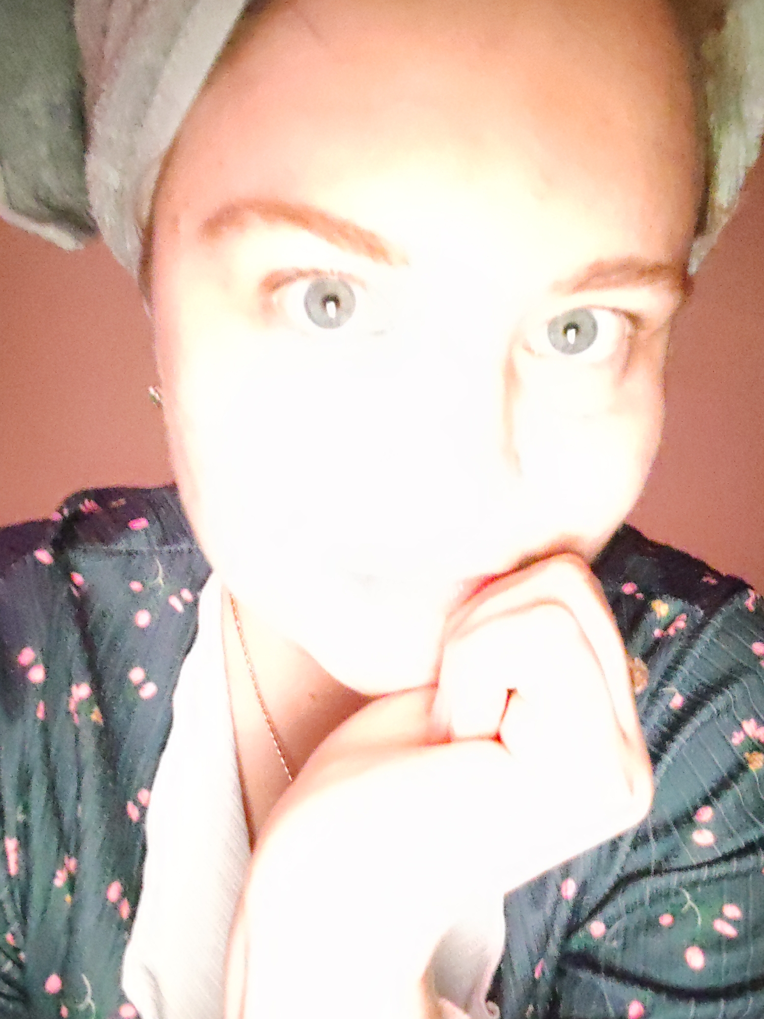 A person with fair skin, light eyes, and auburn eyebrows poses close to the camera. They have a grey towel wrapped around their head. Their right hand is raised to their chin. They are wearing a dark-colored, floral patterned top and a light-colored undershirt. A thin necklace is visible around their neck. The background is a solid, muted red-pink color. The location is not discernible from the image.