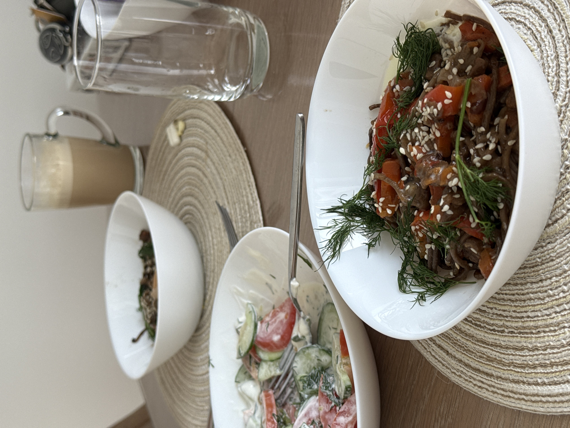 The image shows a table setting with a variety of food items. A glass of light brown liquid, possibly a latte, sits next to an empty glass on a woven placemat. Three white bowls contain different salads; one with tomatoes, cucumbers, and a creamy dressing; another with dark noodles, red peppers, sesame seeds, and dill; and a third, smaller bowl containing a darker salad. A fork rests in the bowl with the tomato and cucumber salad. The setting suggests a meal at home. The city and specific location are unknown.