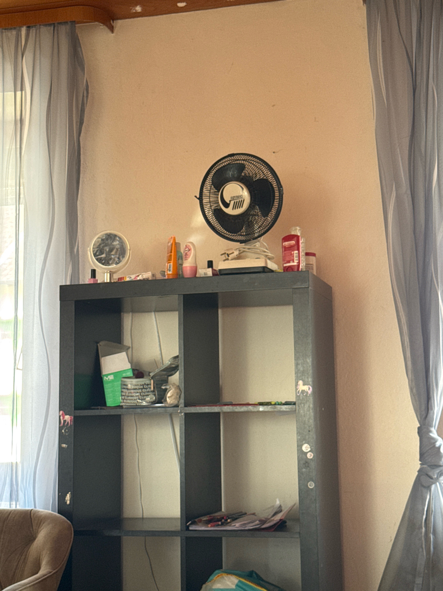 The image captures an indoor scene, likely a room in a residential building located in Überherrn, Germany. The focus is on a dark grey, multi-shelf storage unit against a pale peach-colored wall. On the top shelf, a black oscillating fan is positioned to the right of a small circular mirror. Various small items, including bottles and containers, are scattered on this shelf. Below, the storage unit has several open compartments. The left side of the unit's top shelf displays a green box with white lettering and some crumpled paper. On the left side of the unit, at the mid-level shelf, a small pink and white sticker of a horse is visible. The right side of the unit, at the same shelf level, appears mostly empty. The lower shelves are less visible, but one on the right contains some papers and pencils. The scene is illuminated by natural light coming from a window on the far left, partially obscured by grey curtains. Another set of grey curtains is visible on the right side of the image, tied back with a knot. A portion of a beige upholstered armchair is visible in the bottom left corner of the frame. The overall impression is of a cluttered but lived-in space.