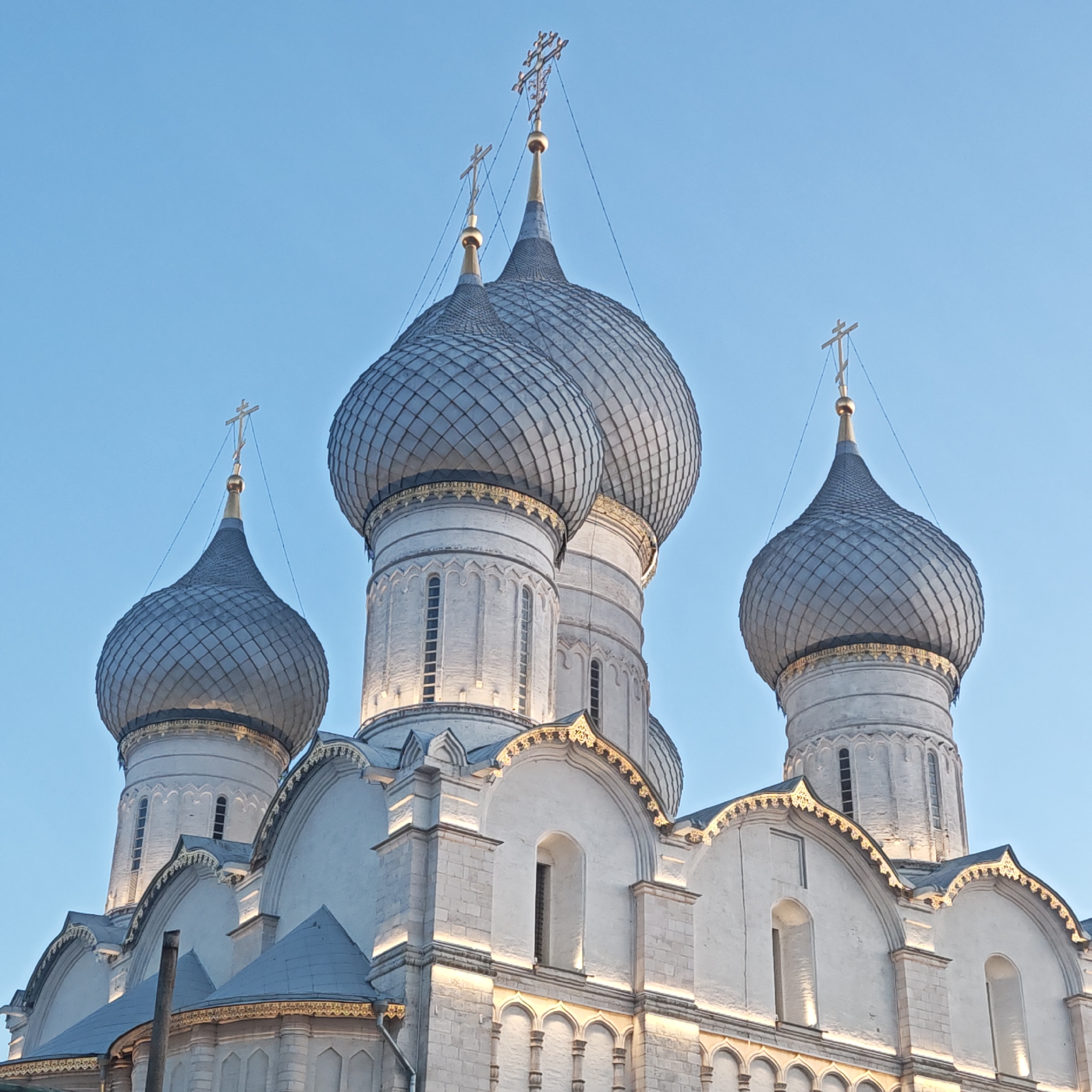 The image shows a portion of the Dormition Cathedral in Rostov, Russia. The architectural style is characterized by white stone walls and silver onion domes, topped with golden crosses. The sky is a clear, pale blue, suggesting either early morning or late afternoon lighting. The cathedral's facade has arched windows and decorative elements. There are no people visible in the frame.