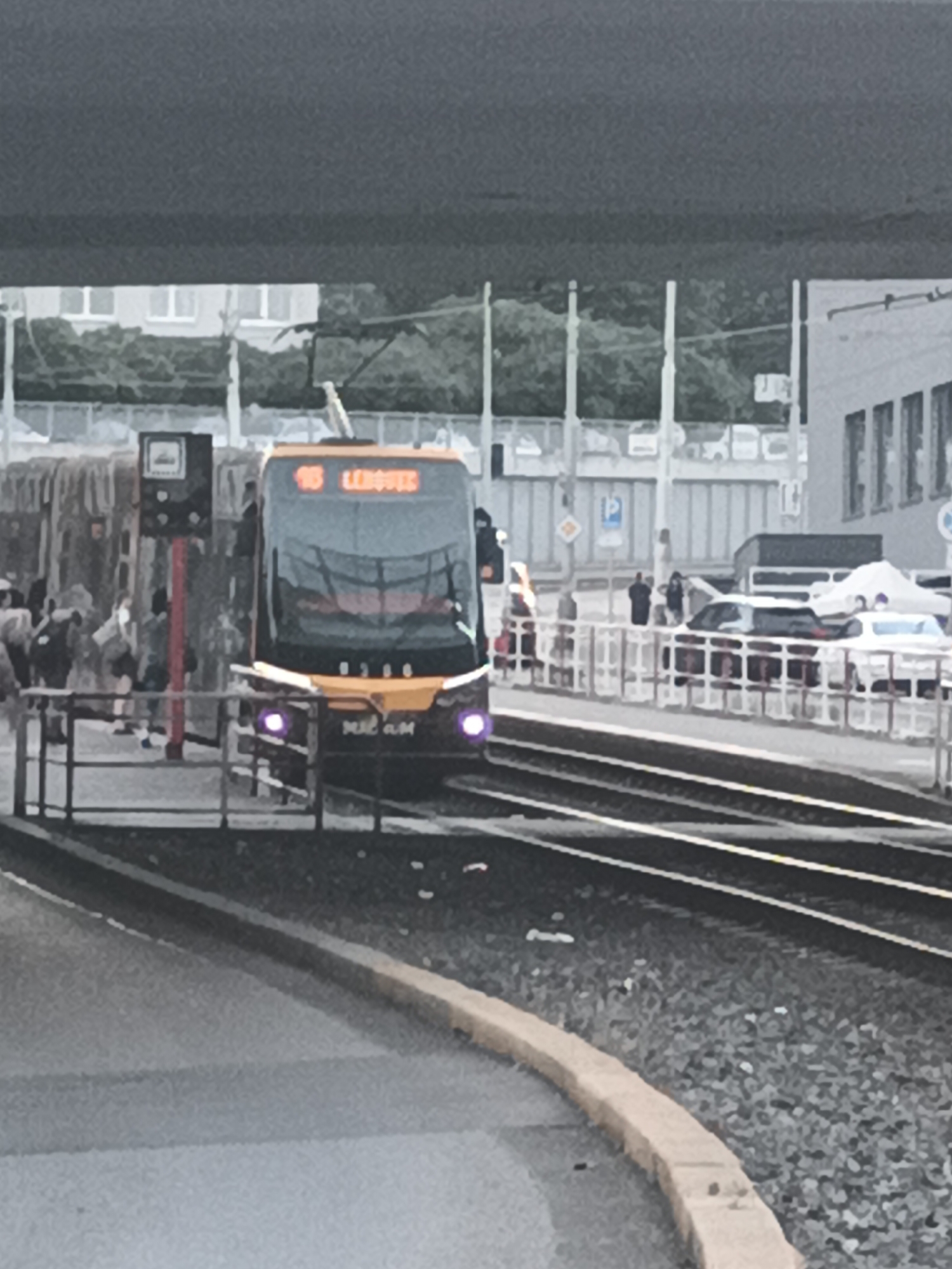 The image shows a tram on tracks in Prague, Czechia. The tram is yellow and has the route number "18" and destination "LEHOVEC" displayed on its front. There are people visible on the platform to the left of the tram, some with backpacks. A traffic signal and a parking sign are also visible. In the background, there are buildings and trees. The overall scene suggests a public transportation hub in an urban environment.
