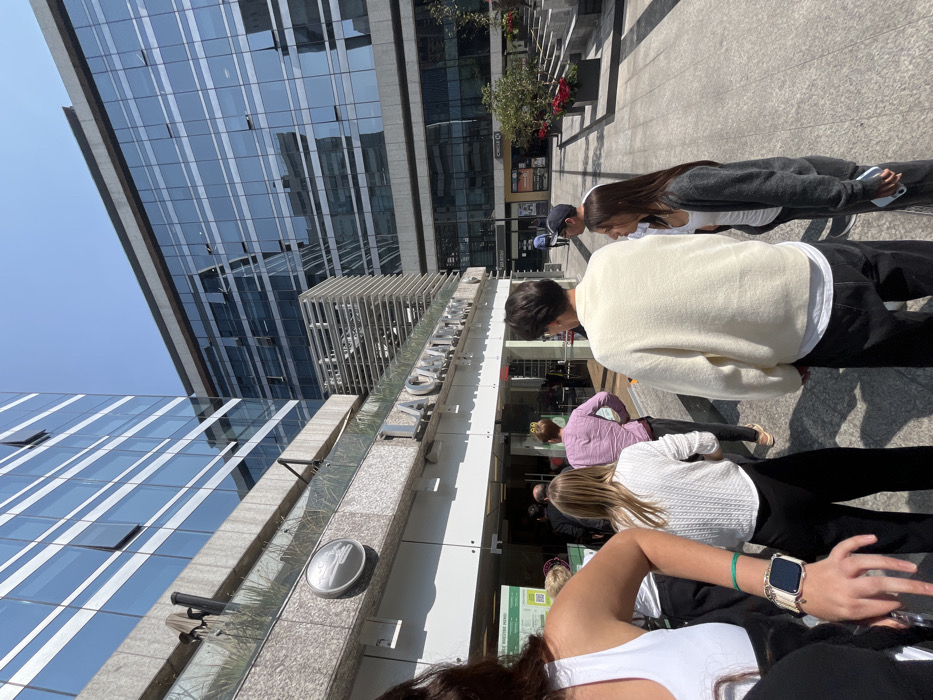 A street-level view captures several pedestrians walking past a modern building with a mirrored glass facade. The building's signage is visible, with large letters. A variety of clothing styles can be seen amongst the people, from casual sweaters and jackets to tank tops, suggesting a temperate climate. The scene is filled with light, casting strong shadows and illuminating the architectural details.
