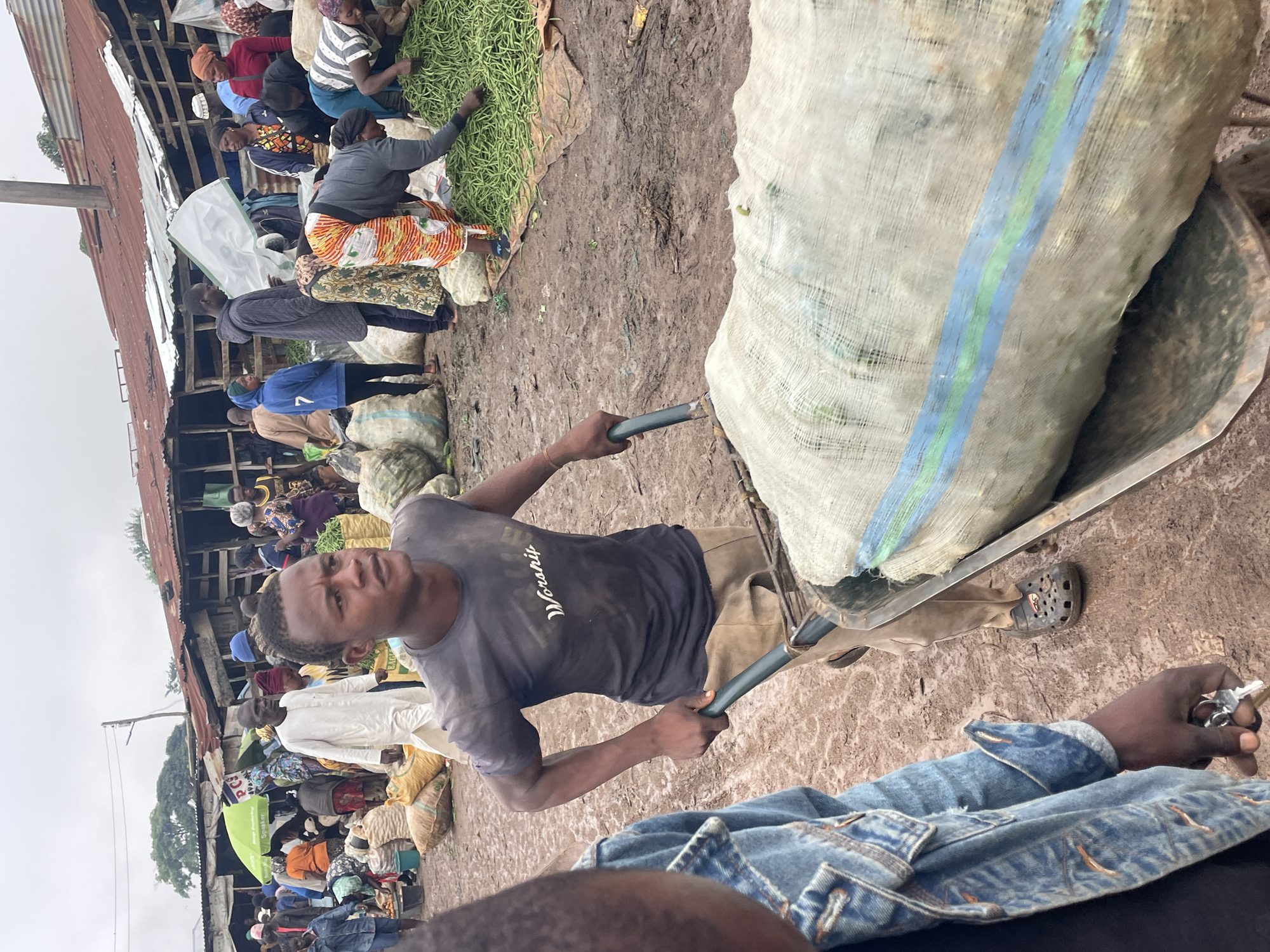 A street market in Gura Top, Nigeria, is depicted on an overcast day. The ground is muddy, suggesting recent rain. A young man in a dark t-shirt and tan pants is pushing a wheelbarrow filled with a large, netted bag containing produce, possibly vegetables, across the uneven terrain. He wears dark Croc-style shoes with a distinct red logo on one. In the foreground, the arm and shoulder of another person wearing a denim jacket are visible. In the background, numerous people are engaged in market activities, with stalls selling goods under corrugated metal roofs. Some individuals are handling large piles of green beans, and other produce is visible in sacks and baskets. The atmosphere appears busy and dynamic, typical of a local market.