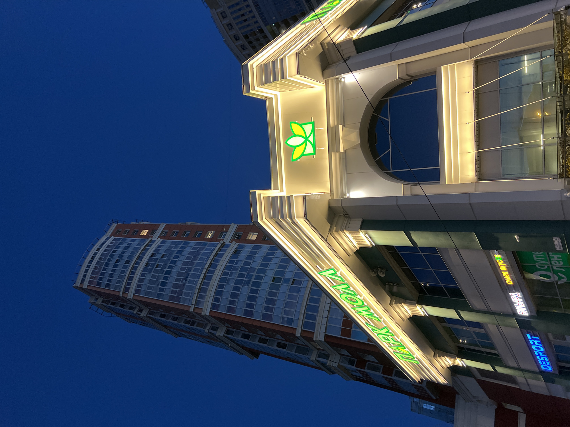 A low-angle, wide shot captures a modern multi-story building at dusk in an urban environment. The building features a prominent neon sign with green Cyrillic text that reads "ПАРК МОЛЛ" (PARK MALL) illuminated against a dark blue sky. Above the sign, a green floral logo is also lit. The building's facade is white with decorative columns and horizontal yellow lighting. To the left, a taller, curved skyscraper with numerous windows is visible, its brick exterior contrasting with the illuminated lower building. Further to the right, another building with a decorative cornice and illuminated windows is partially visible. The image is taken at ground level, looking upwards, with the street-level entrance area of the "PARK MALL" featuring shops with neon signage, including "DESPORT" in blue.