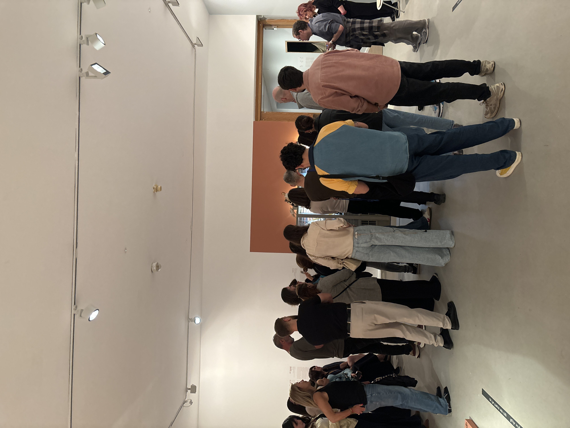 The image captures an indoor scene, likely an art gallery or exhibition space, filled with a diverse group of people observing exhibits. The perspective is from behind and slightly above the crowd, looking towards the front of the room.
**Subjects:** There are at least 20-30 individuals, primarily young to middle-aged adults, dressed in casual attire including jeans, t-shirts, sweaters, and sneakers. Their backs are mostly to the camera, and many are leaning forward or looking down, indicating engagement with displays or artworks positioned at eye level or slightly below.
**Activity:** The people are attending an exhibition or gallery viewing. They are standing in rows or clustered groups, suggesting they are moving through a designated viewing area, attentively observing unseen items on the walls or pedestals.
**Setting:** The room features light-colored walls, predominantly off-white, with a section of the back wall painted a contrasting warm orange-brown hue. The floor is a light grey concrete or polished surface. Track lighting with small, bright spotlights is mounted on the walls and ceiling, illuminating the space, particularly the display areas. A wooden doorway is visible in the background, leading to another room.
**Notable details:** The lighting is bright and focused, characteristic of an exhibition setting. The atmosphere appears quiet and contemplative, with attendees focused on the exhibits. On the grey floor in the foreground, there is visible text. Part of it is in English, reading "ON OR LIKELY THE UMS". Below this, there is text in Hebrew, which translates to "or agreed by", confirming the location context of Tel-Aviv, Israel.
