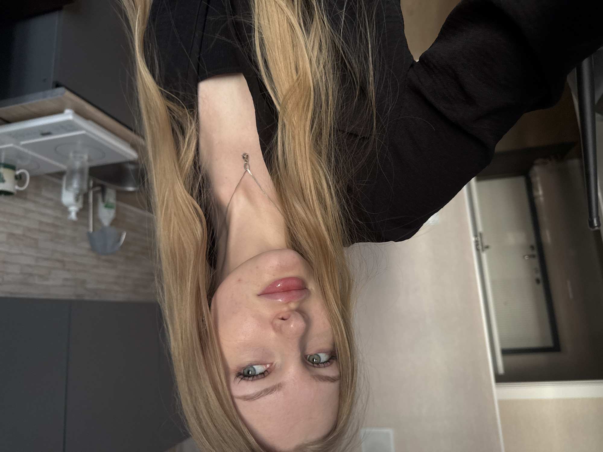 A woman with long, blonde hair is facing the camera. She is wearing a black top and a silver necklace. The background shows part of a kitchen, including a stove top, cabinets, and a door. It is unknown whether this photo was taken in Perm, Russia.