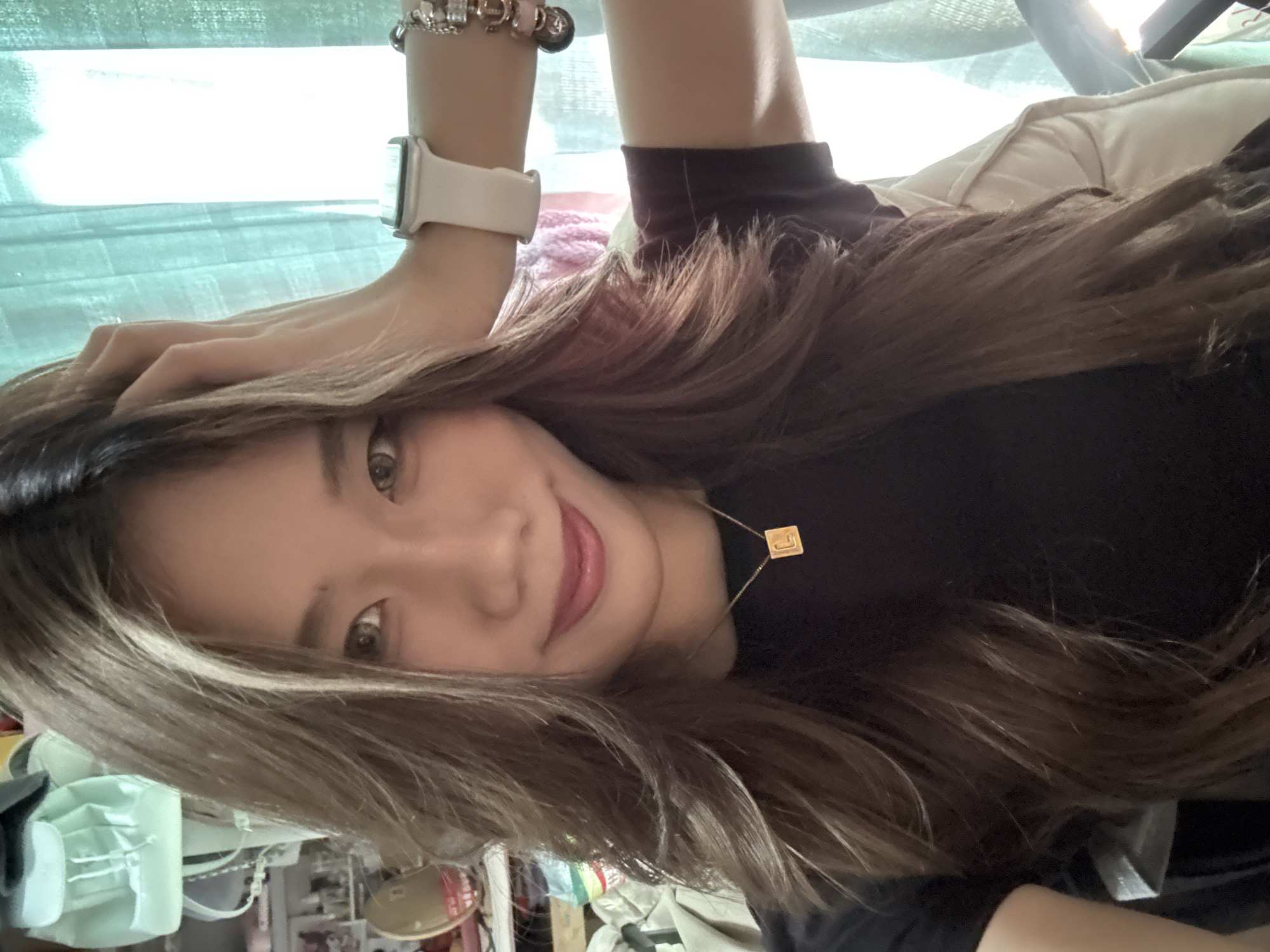 A young woman with long brown hair is lying on her back, smiling at the camera. She is wearing a black shirt and a gold necklace with a square pendant. Her left hand is resting on her hair, and she is wearing a white smartwatch and a bracelet with charms. The background appears to be a cluttered room with a window. The location cannot be confirmed as Nan'an District, China without additional information.