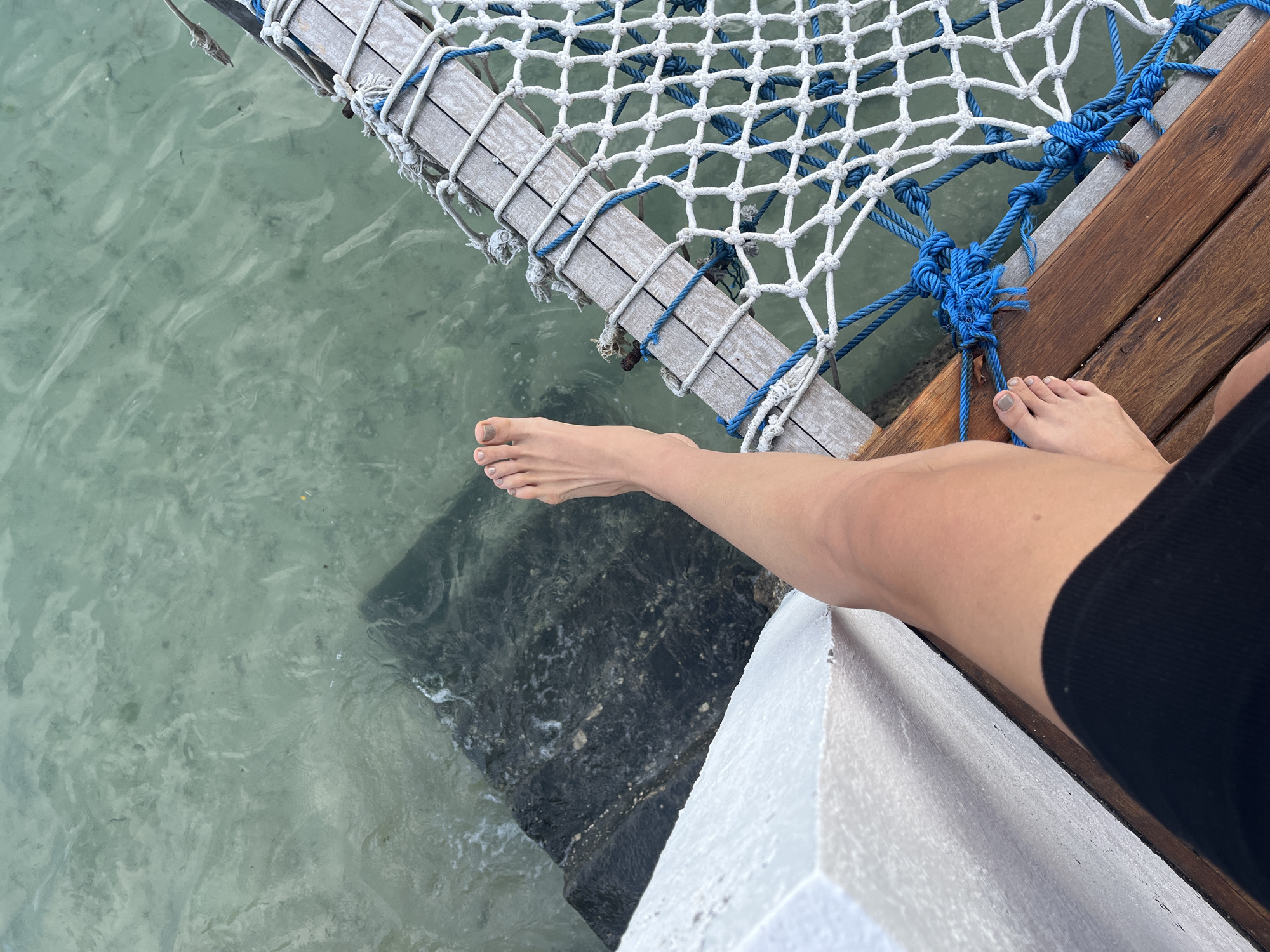 A first-person perspective shot shows a bare left foot with gray nail polish, and a portion of a tanned leg extending from the bottom of the frame upwards, positioned on a wooden deck. To the right of the leg and foot, a white and blue netted structure, possibly a hammock or a safety net, is attached to wooden beams. The water below is clear and greenish, revealing dark rocks and sandy patches. A white painted structure is visible on the left side of the image. The scene appears to be a waterfront area, likely a resort or a pier, in Nusa Penida, Indonesia.
