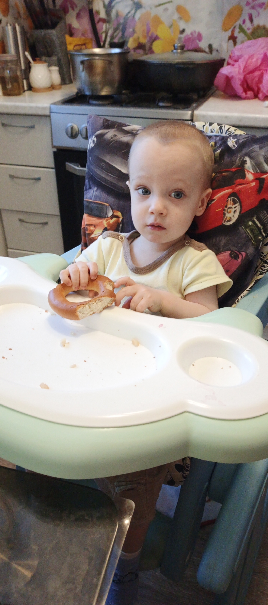 A young child with light hair and blue eyes, wearing a light yellow top with brown trim, is seated in a high chair in a domestic kitchen in Chaikovsky, Russia. The child holds a partially eaten bagel in both hands, looking directly at the viewer. Bread crumbs are scattered on the white tray of the high chair, which also features a light green base and a backrest patterned with images of sports cars in red, orange, and black. In the background, a kitchen counter is visible, with a floral wallpaper displaying yellow, purple, and pink patterns above it. A silver gas stove with an oven is centered on the counter, holding a metal pot and a darker pot, both with lids, on its burners. To the left, a glass jar, a white sugar bowl, and a creamer are on the countertop. A crumpled pink plastic bag rests on the counter to the right of the stove. Light grey kitchen cabinets with horizontal handles are visible below the countertop.