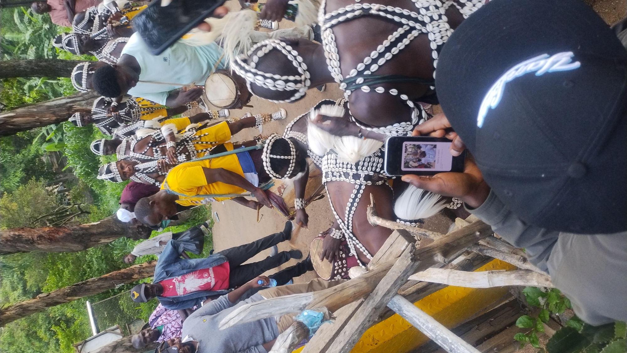 The image depicts an outdoor gathering in Jos, Nigeria, likely a cultural event or ceremony. A group of men, some adorned in traditional attire featuring shell necklaces and headdresses, are engaged in an activity that appears to involve drumming and possibly music. One individual is playing a drum, while others seem to be participating in the rhythm.
In the foreground, a person is holding a smartphone, capturing the scene. The phone screen displays a smaller image of individuals, potentially related to the event or a broader context. The background consists of trees and vegetation, suggesting a natural or semi-natural environment within the city. The ground is dirt, typical of many outdoor spaces in Nigeria. The overall atmosphere appears lively and communal.