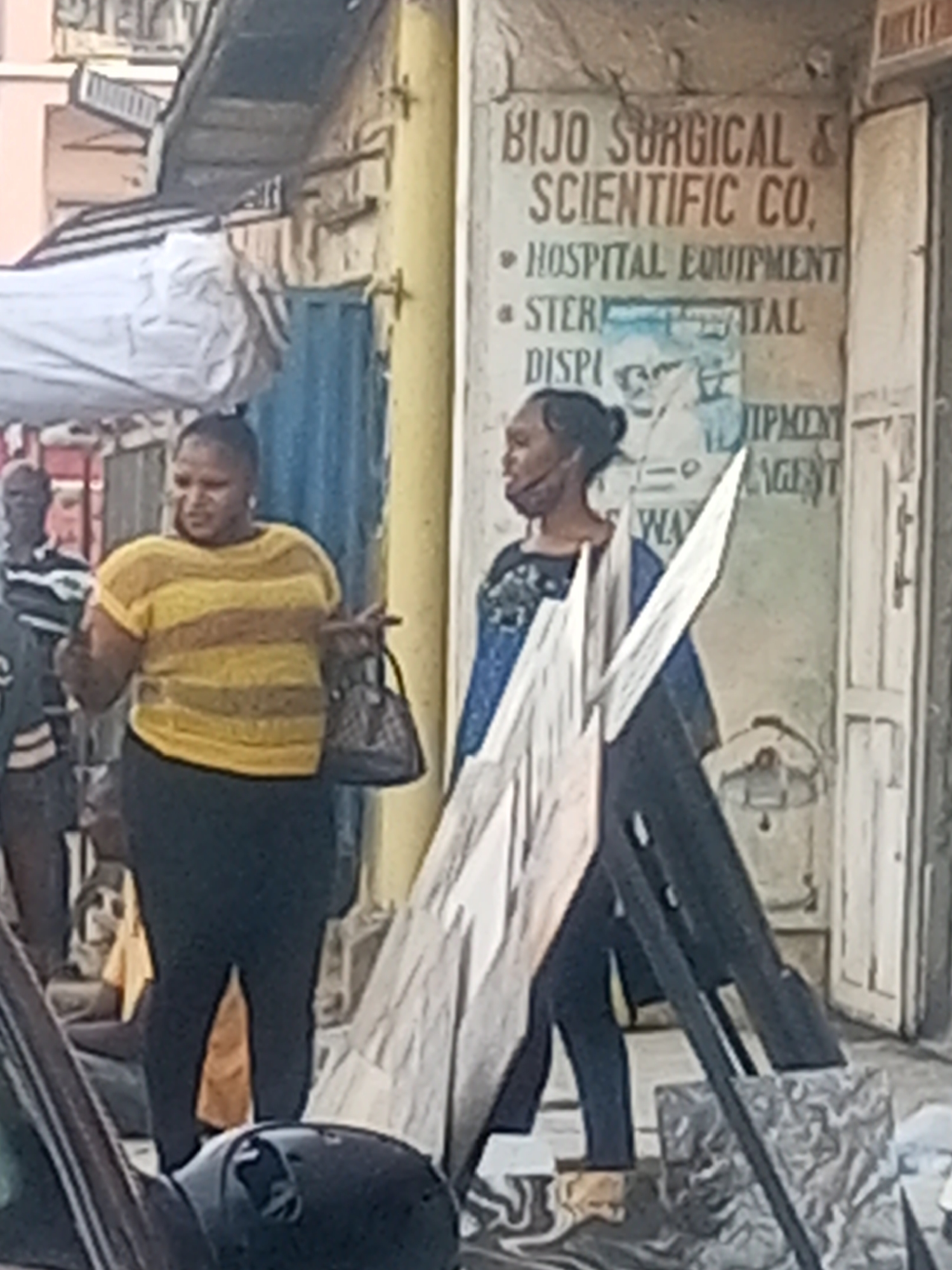 The image depicts a street scene in Maiburiji, Nigeria, with two women standing in front of a business called "BIJO Surgical & Scientific Co." One woman is wearing a yellow and brown striped shirt and black pants, holding a purse, and gesturing with her hands. The other woman, standing closer to the building, wears a blue top and dark pants, with an object obstructing part of her lower body. A man is partially visible in the background, wearing a striped shirt. A car's side mirror is visible in the foreground. A sign for hospital equipment, sterile disposal, and equipment agent can be seen on the building wall. There is also some makeshift construction material.