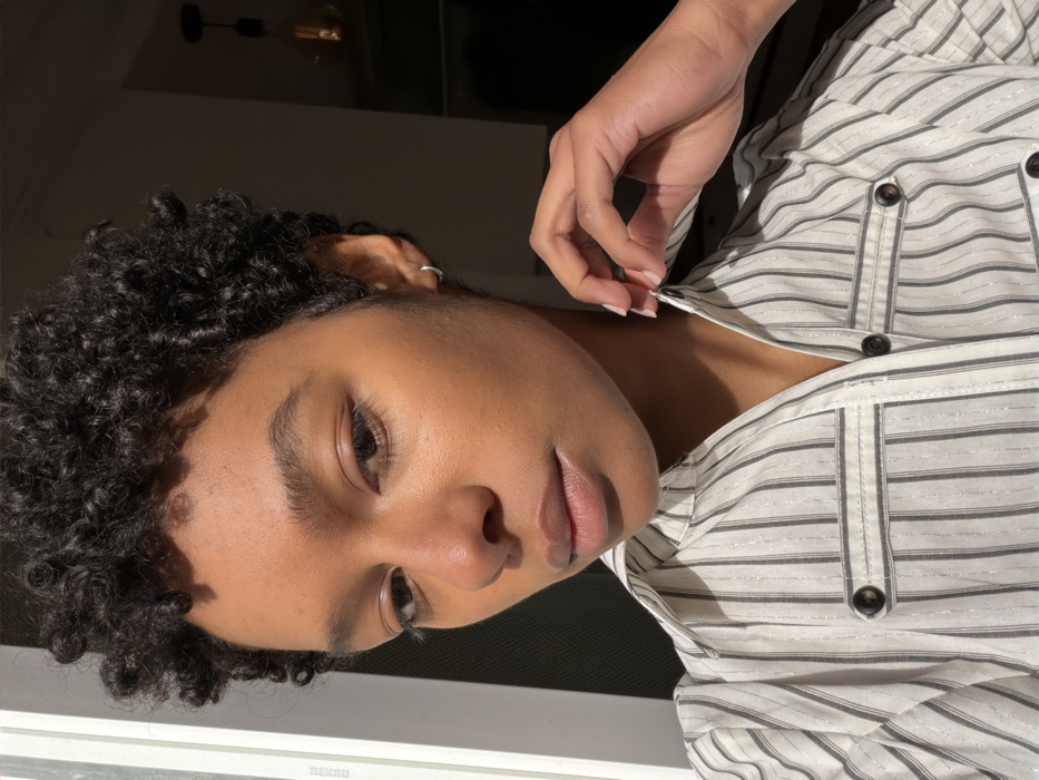 A close-up, chest-up portrait features an individual with medium-brown skin and short, dark, tightly curled hair. The person wears a light-colored, vertically striped collared shirt with thin dark grey or black stripes and subtle metallic threads. The shirt includes two decorative breast pockets with dark, round buttons. A small silver hoop earring is visible in the subject's left earlobe. The subject's right hand, with manicured nails, gently touches the shirt's collar near the neck. The individual gazes directly forward. Strong natural light illuminates the left side of the subject's face and body, originating from an unseen window to the left. The background is dim, revealing a dark wall or interior space on the right, where a pendant light fixture with an exposed Edison-style bulb hangs. The specific location within Paris, France, cannot be determined from the visual evidence provided.