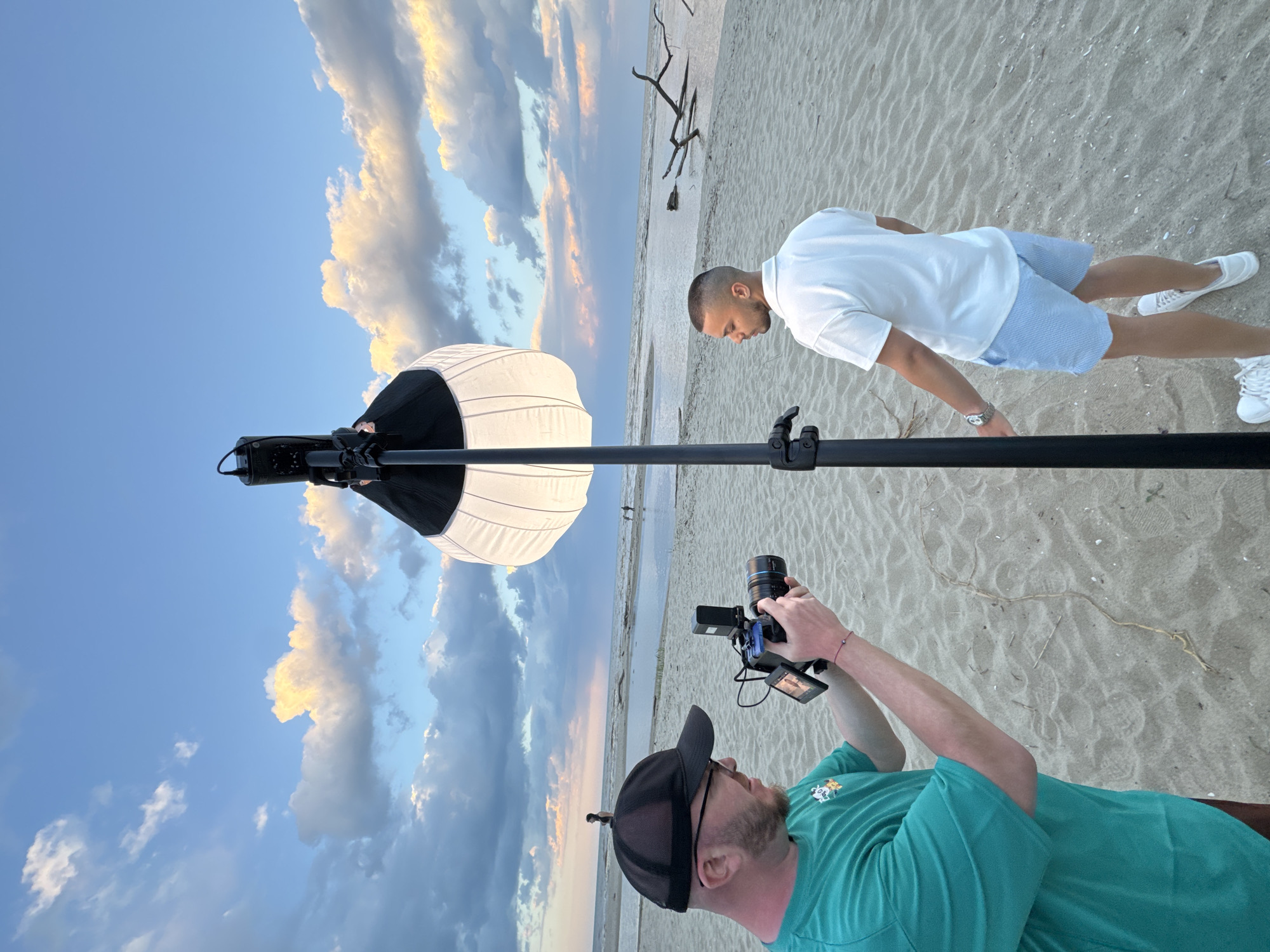 Two men are on a beach. One man in a green t-shirt and black baseball cap is holding a camera and filming the other man. The man being filmed is wearing a white polo shirt, blue shorts, and white sneakers. A large white light diffuser is positioned on a stand between the photographer and the subject. The beach is sandy, and there are some branches lying on the sand in the background. The sky is cloudy with patches of blue. The location is likely Novo Oriahovo, Bulgaria, based on the presence of a beach.