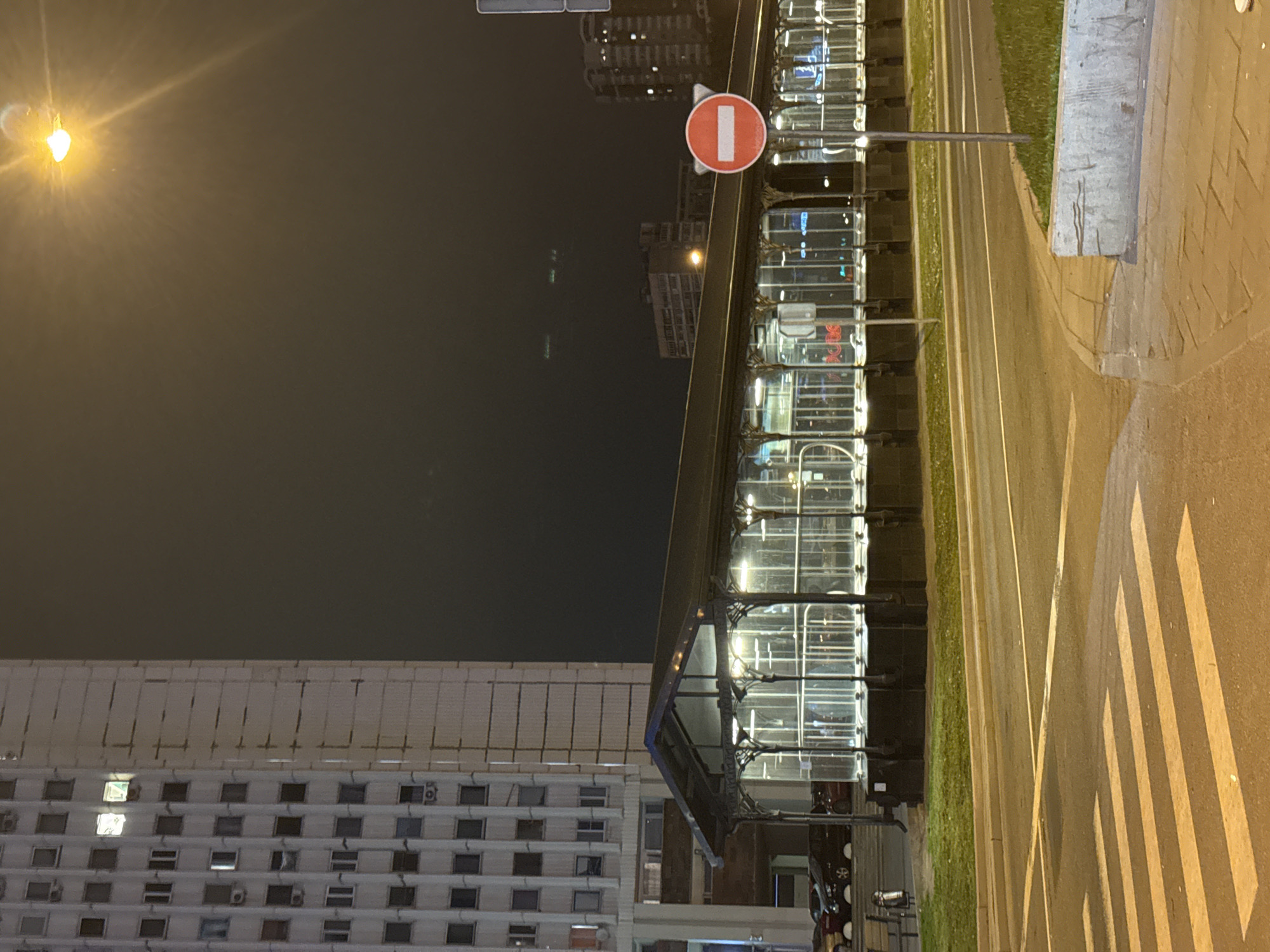 This image, taken at night in Moscow, Russia, depicts a modern, glass-encased bus station or transit hub alongside a multi-lane road. The lighting is primarily artificial from streetlights and interior illumination of the structure. The environment shows a combination of urban infrastructure (buildings, road, crosswalk), and minimal greenery. There is a "no entry" road sign. Adjacent to the transit hub is a tall, light-colored apartment building with many windows, some with lights on. No individuals are clearly visible.