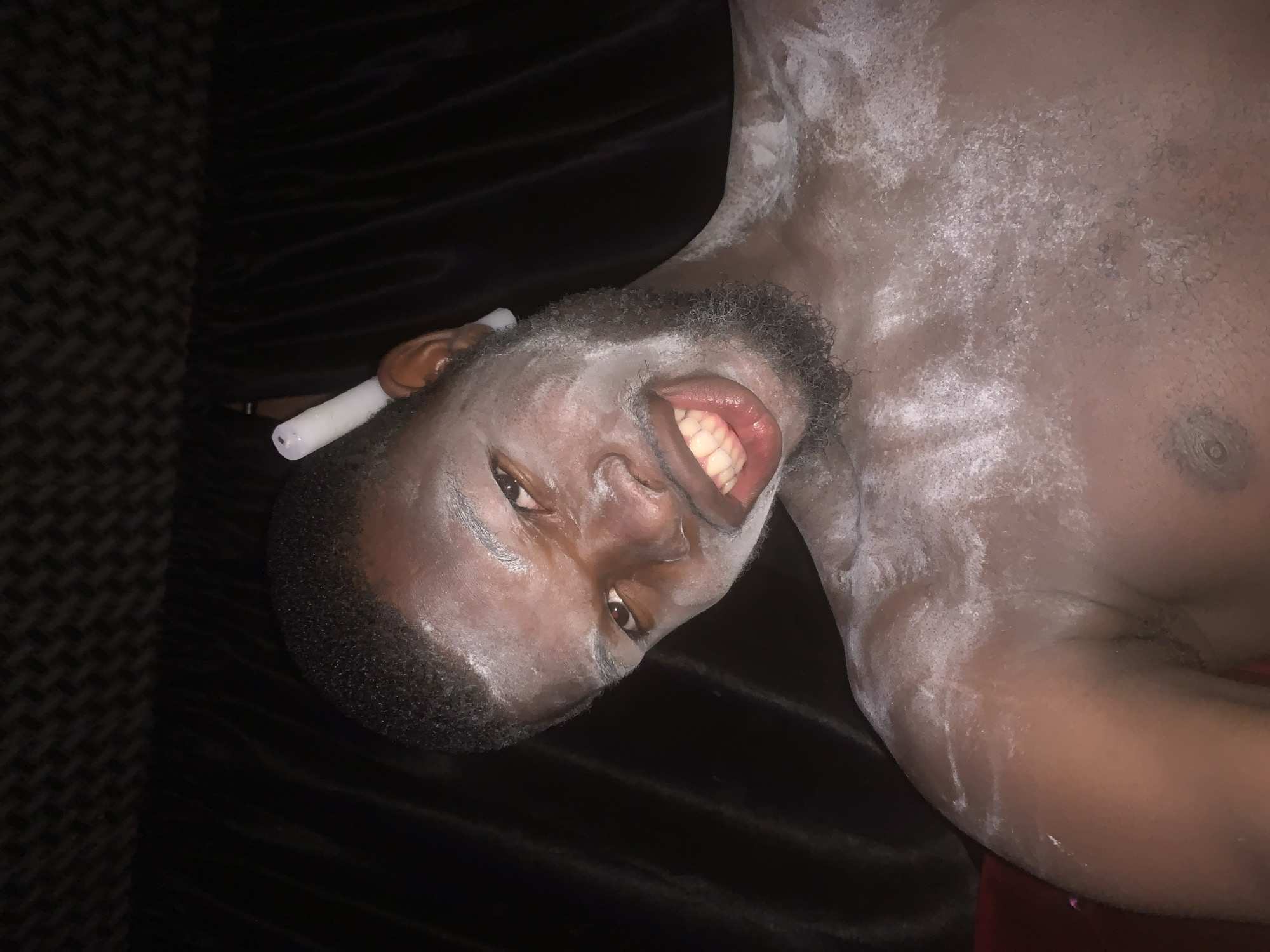 An image, captured in a low-light indoor setting, displays a dark-skinned individual, likely male, from the chest up. The person is positioned sideways, facing the camera with their head tilted, and their body largely covered in a fine white powdery substance, reminiscent of talcum powder, on their face, neck, chest, and shoulders.
The individual has short, dark hair and a beard. Their mouth is open wide, revealing their teeth in what appears to be a broad smile or playful expression, and they are looking directly into the camera. A white, cylindrical object, resembling a vape pen or e-cigarette, is held by their left hand near their left ear. The background is predominantly dark, suggesting dark fabric or bedding, with a patterned, possibly carpeted, surface visible on the left side of the frame. There are no visible location cues within the image itself, though the context indicates it was taken in Onitsha, Nigeria. No text is visible in the image.