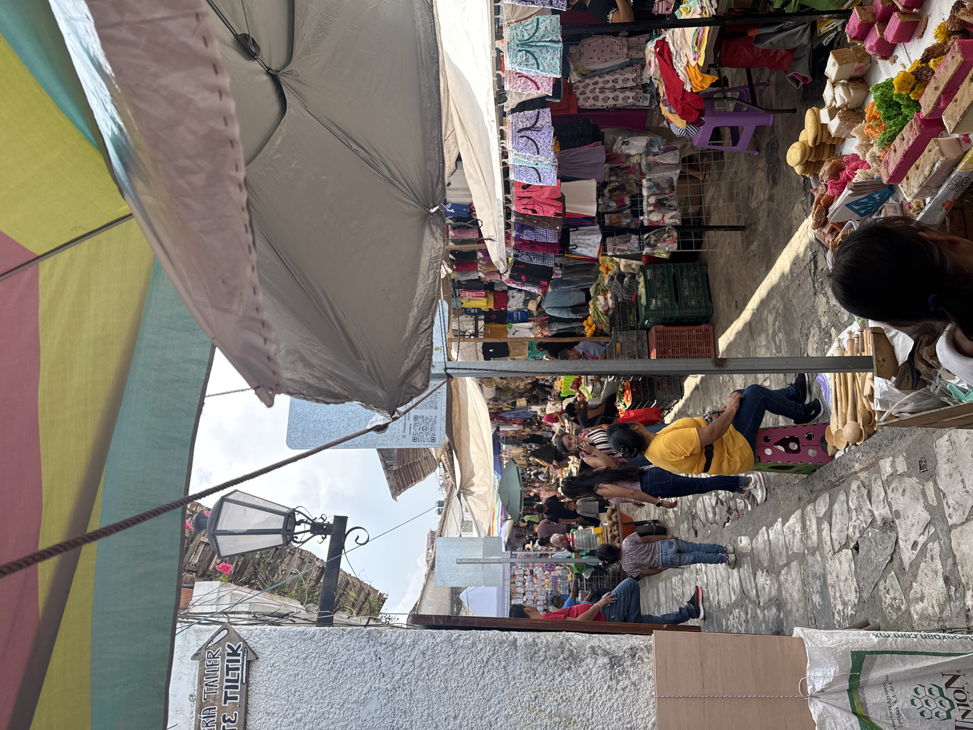 Here's a description of the image:
The image shows a vibrant street scene at the outdoor market in the town of Cuetzalan, Mexico. The market is held on a narrow, cobblestone street. People are browsing the various stalls lining the street. The stalls are selling a variety of goods, including clothing, food items like candy and baked goods, and other merchandise. Many of the stalls are covered with large umbrellas or awnings to provide shade from the sun.
The view is looking down the street, showing a number of vendors seated along the pavement, with customers walking past. Several people are visible in the foreground and background. A lamp post and building are visible to the left.