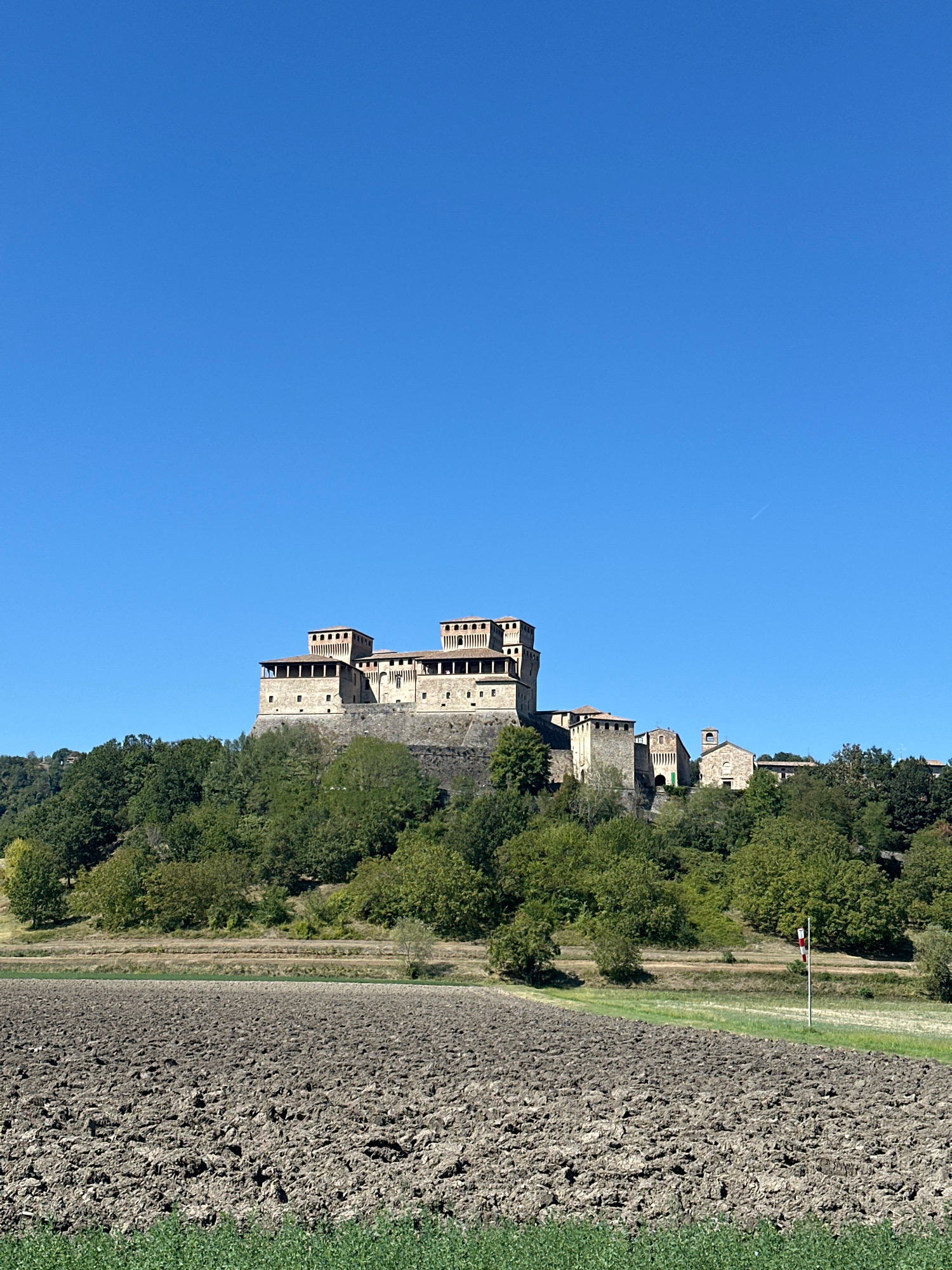 The image depicts a medieval castle complex, likely an Italian fortress, situated atop a verdant hill under a clear, bright blue sky. In the foreground, a dark, freshly plowed agricultural field occupies the lower half of the frame. A strip of green vegetation borders the bottom edge of the field. Beyond the field, a band of trees and shrubs ascends the hillside towards the castle. The castle itself is a substantial stone structure with multiple towers and crenellated battlements, exhibiting a weathered, historic appearance. Adjacent to the main castle structure, a smaller, simpler stone building with a pitched roof, possibly a church or chapel, is visible. A white flagpole with red and white markings stands in the middle ground to the right, in front of the hill and field. The overall scene suggests a rural setting in a historically significant location, possibly the Italian countryside near Langhirano.