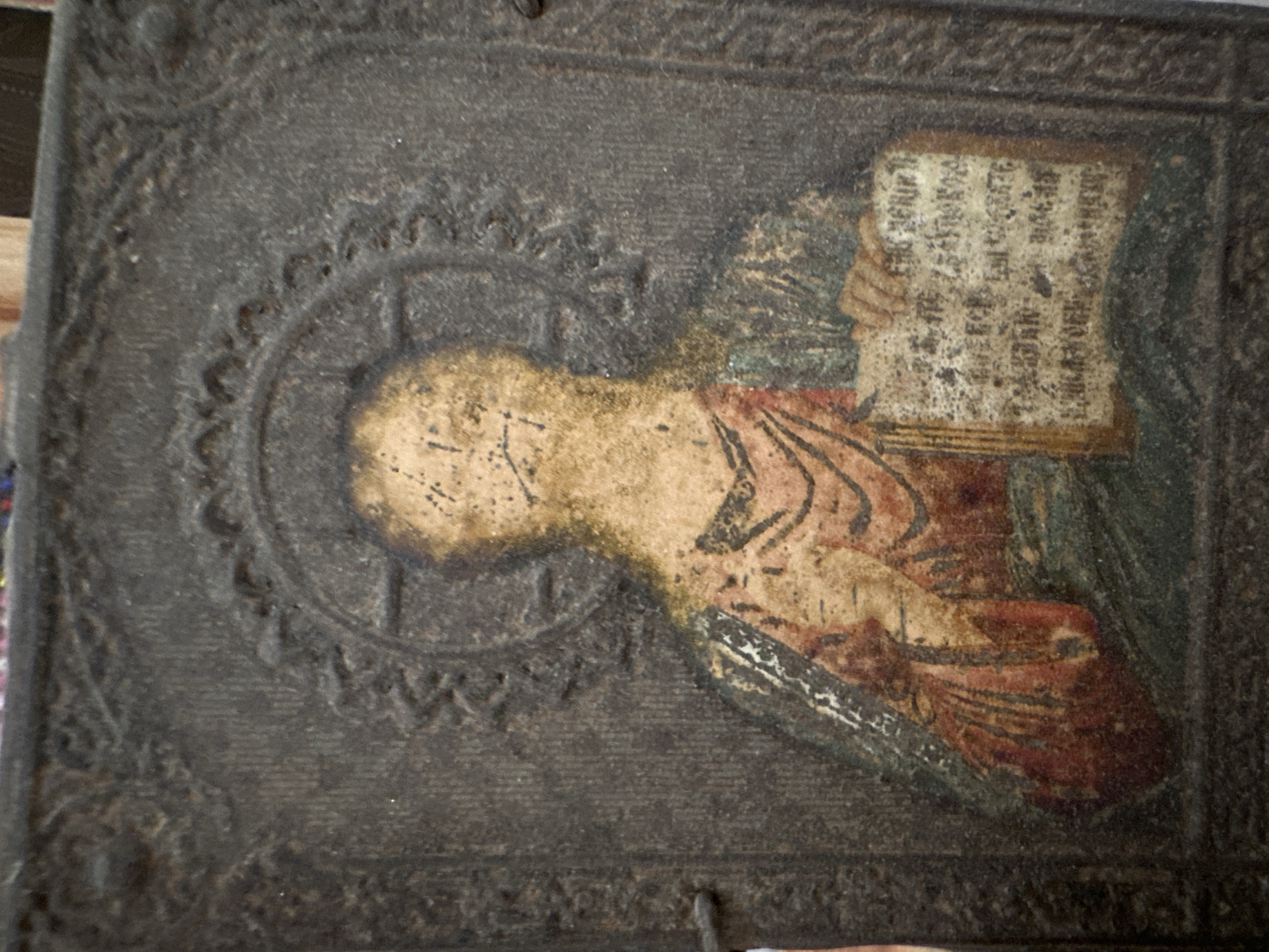The image shows an aged religious icon, possibly Orthodox, depicting a figure with a halo holding a book. The icon has a dark, weathered appearance with visible wear and deterioration of the paint. The figure is wearing robes and holding a book with script, though the script is heavily degraded. The icon is encased in what appears to be a dark, decorative border. The location in Sayansk, Russia, is based on external information, and does not appear in the image itself.