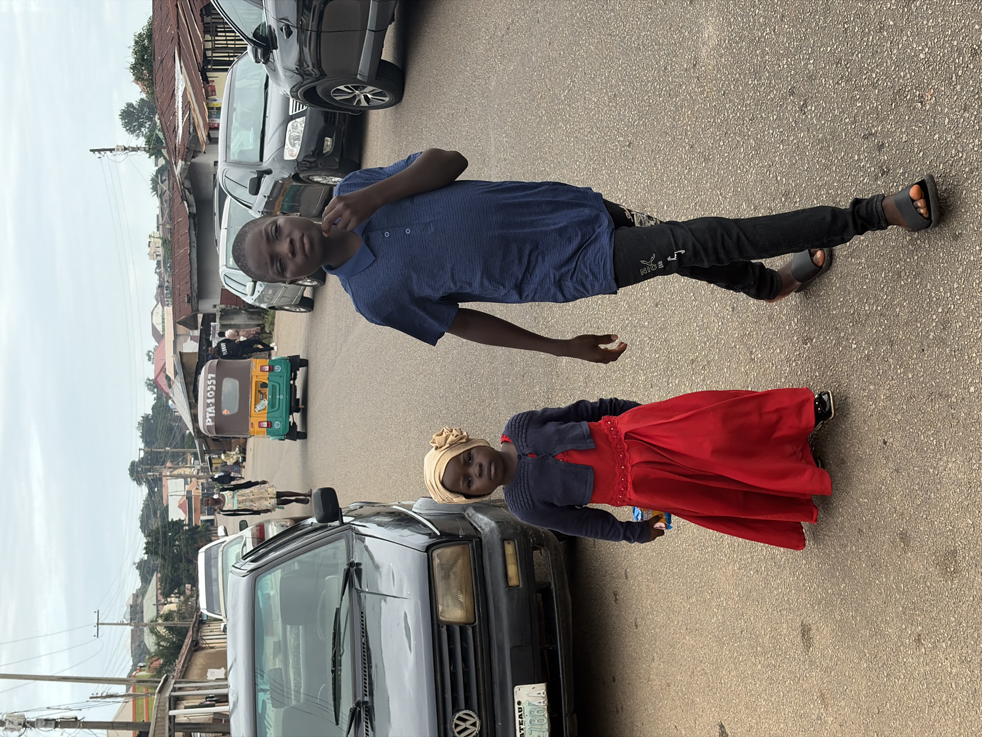 This image captures a street scene in Jos, Nigeria. In the foreground, a young boy in a blue collared shirt and black pants walks towards the camera, his hand touching his chin. To his left, a young girl in a red dress and a tan headscarf with a floral embellishment stands beside a dark Volkswagen car. She is holding a small packet. Behind them, other vehicles, including a colorful auto-rickshaw with "PTA-10337" visible on its side, are parked or moving along the asphalt road. Buildings with corrugated iron roofs line the street, suggesting a typical urban or semi-urban environment in Jos. The sky is overcast, contributing to the diffused lighting of the scene.