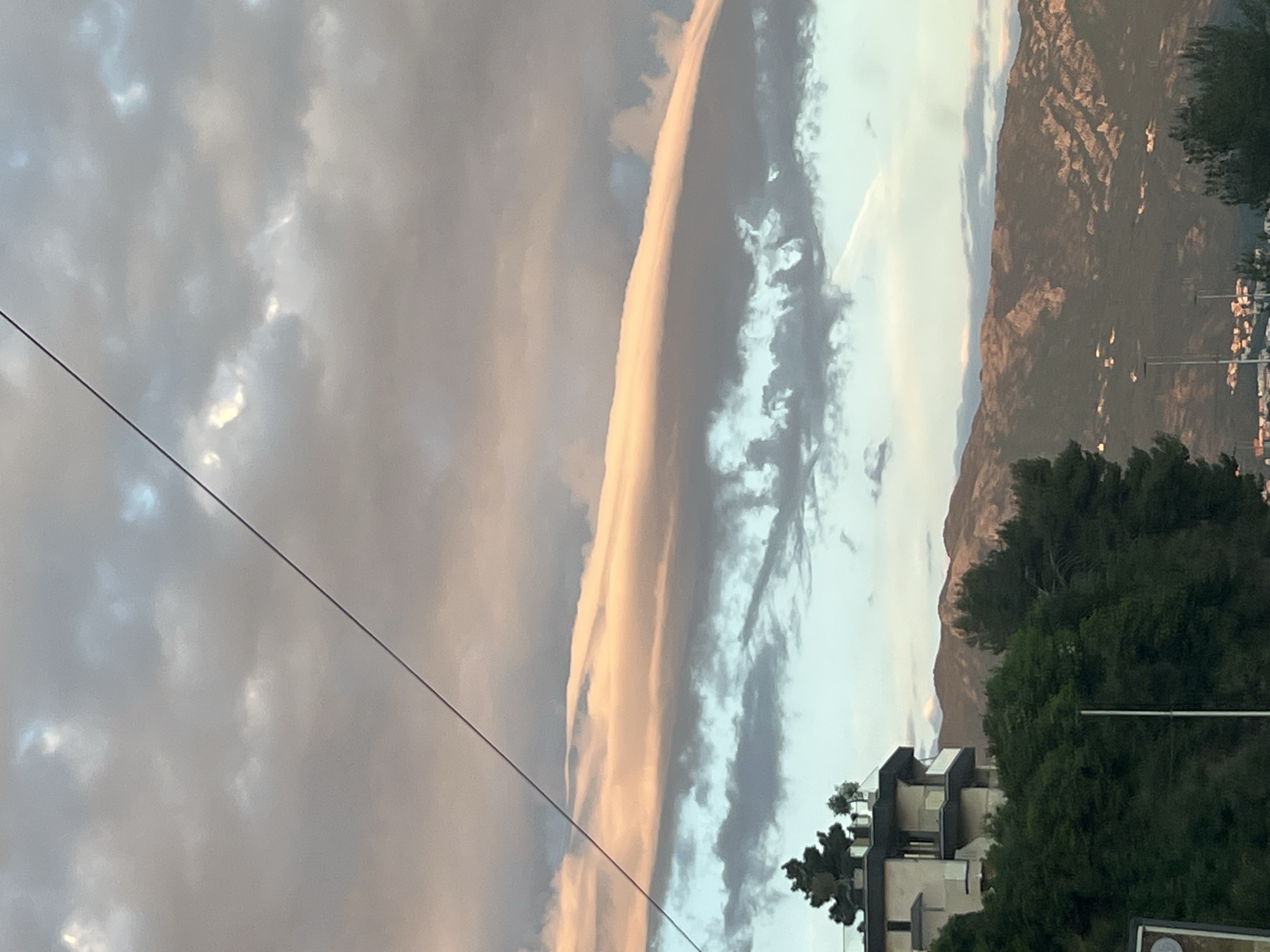 The image captures a landscape view of a mountainous coastal area, consistent with Budva, Montenegro, likely during late afternoon or early evening. The upper portion of the frame is dominated by a cloudy sky. A prominent, elongated cloud stretches horizontally across the middle of the sky, illuminated with warm golden and pink light, suggesting the sun is low on the horizon. To its left, darker grey clouds are visible, while to its right, lighter blue sky with scattered wispy clouds can be seen. A thin, dark power line diagonally traverses the upper-left section of the sky.
Below the sky, a rugged mountain range forms the midground, displaying textured slopes. Some areas of the mountain appear illuminated, enhancing their contours. In the foreground and lower right, dense green trees partially obscure modern buildings, which appear to be multi-story structures with distinct architectural features like balconies. Another power line or cable is visible extending diagonally from the lower left. There are no people visible, nor is there any discernible text. The scene conveys a tranquil, scenic environment with a mix of natural elements and urban development under a partly cloudy sky with soft, warm lighting.