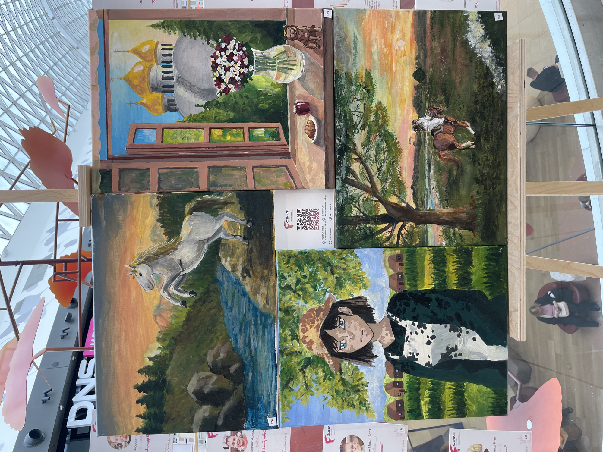 This media file displays a collection of four paintings, likely an art exhibition or a display of student work, as indicated by the presence of identification labels with names and numbers (e.g., "22A", "23A", "24A", "17г", "29г"). The overall setting appears to be indoors, possibly a public space like a mall or gallery, given the modern architectural elements visible in the background, such as a large, geodesic-like glass ceiling and metallic structures.
The paintings themselves depict various subjects:
* The top-left painting shows a view through a window, revealing golden-domed buildings with a bouquet of flowers and a small pastry on the windowsill.
* The top-right painting depicts a rural landscape at sunset, with a horse and rider in motion across a field. The warm hues of the sky suggest it's either sunrise or sunset.
* The bottom-left painting features a dynamic scene of a white horse leaping over a river, set against a backdrop of mountains and a colorful sky.
* The bottom-right painting is a portrait of a young person with distinctive blue eyes, wearing a patterned hat and jacket, with a stylized rural village in the background.
The presence of text in Russian, including names like "Зубова Ю", "Леонард Б", "Андр", and numbers possibly referring to participant or artwork IDs, along with a phone number "89655700519", suggests a Russian context, specifically Perm, Russia, as provided. The labels also mention "DNS", which is a Russian electronics retailer, further hinting at a commercial or public space. There are also cutouts of stylized pink birds or figures, adding a decorative element to the display. The overall impression is one of a vibrant display of artistic talent, likely showcasing local artists or students.