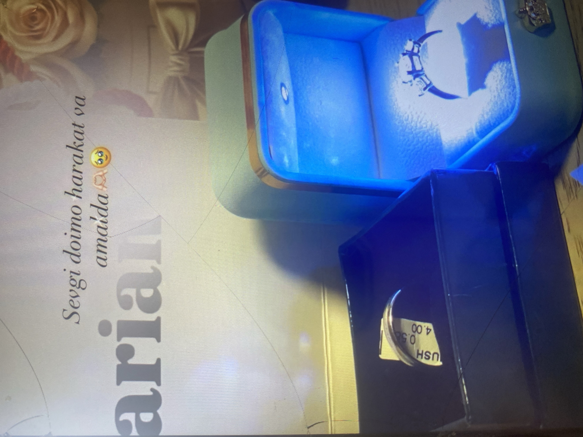 The image depicts two jewelry boxes, one open and illuminated with blue light, showcasing a diamond ring. The second box is closed and dark, with a tag visible on the side of a ring inside. The background features text that appears to be "Sevgi doimo harakat va amaida" followed by emojis, and a large blurred word "arian". A bouquet of flowers is also visible in the background. There is no discernible information about the city of Arbagish, Uzbekistan, in the image.