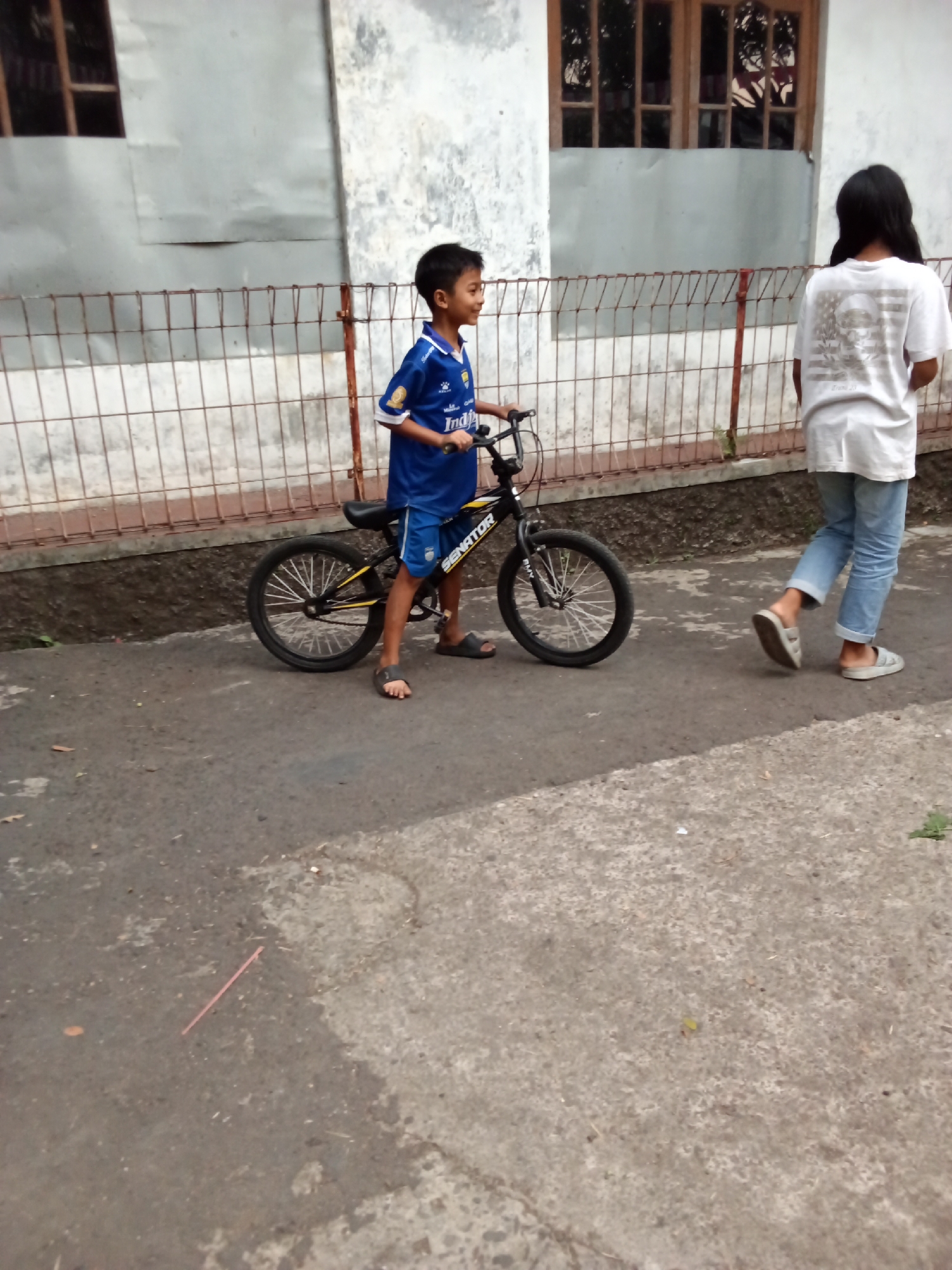A young boy, appearing to be around 8-10 years old, stands on a bicycle. He is wearing a blue soccer jersey with white trim and blue shorts. His feet are clad in dark sandals. The bicycle is black and yellow, with the brand name "SENATOR" visible on the frame. The boy is looking to his right, with a slight smile. To the right of the boy, a young girl, appearing to be around 10-12 years old, walks away from the camera. She has long black hair and is wearing a white t-shirt with a graphic design on the back and blue jeans. She is wearing light-colored sandals. They are on a paved surface, likely a street or sidewalk, in what appears to be a residential area. In the background, there is a building with windows and a fence. The overall setting suggests a casual outdoor environment in a city. Given the details of the architecture, typical of many Indonesian cities, and the clothing styles, this scene could plausibly be located in the city of Bandung, Indonesia.