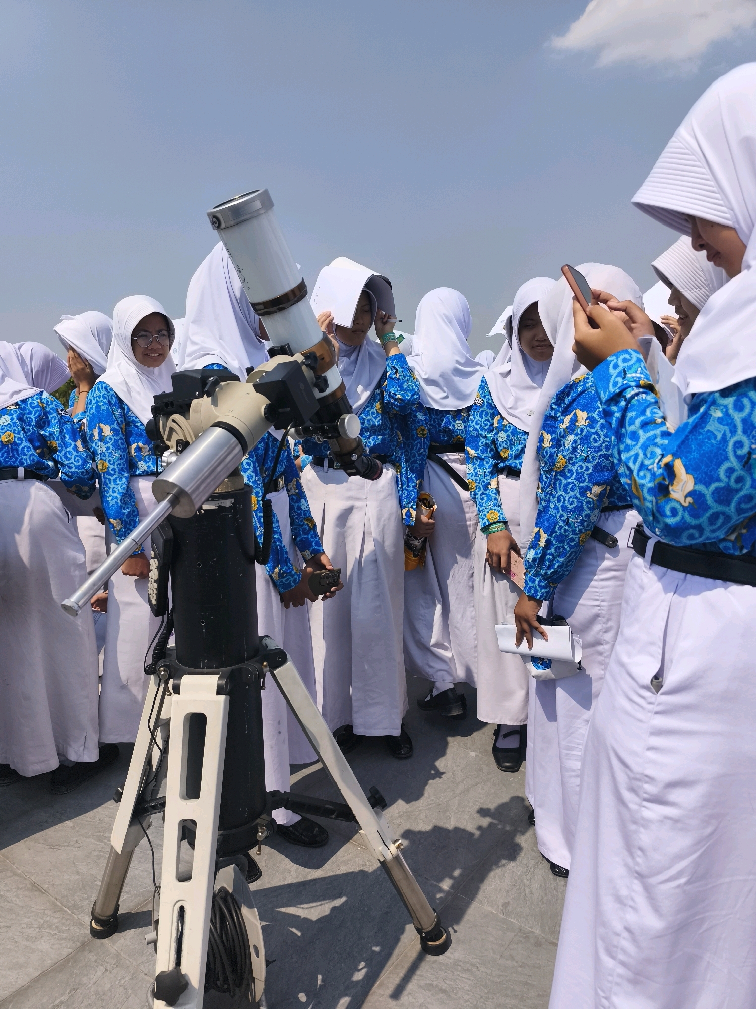 A group of Muslim students in white hijabs and blue patterned uniforms stand around a telescope on a tripod, outdoors. The students appear to be looking at the telescope or at their phones. One student on the right is holding a phone up, possibly to take a picture. The sky is visible in the background, with some clouds. The ground appears to be paved with light-colored tiles. The scene is well-lit, suggesting it is daytime. The blue uniforms have a repeating pattern of what appear to be stylized birds or mythical creatures. The telescope is a long, white cylinder with various attachments and knobs. The students are positioned in a way that suggests they are observing something through the telescope. The presence of the telescope and the clear sky suggest an astronomical observation event. The clothing of the students, particularly the hijabs and uniforms, may indicate a school event or activity. Without further context, it is difficult to ascertain the exact location within Semarang, Indonesia.