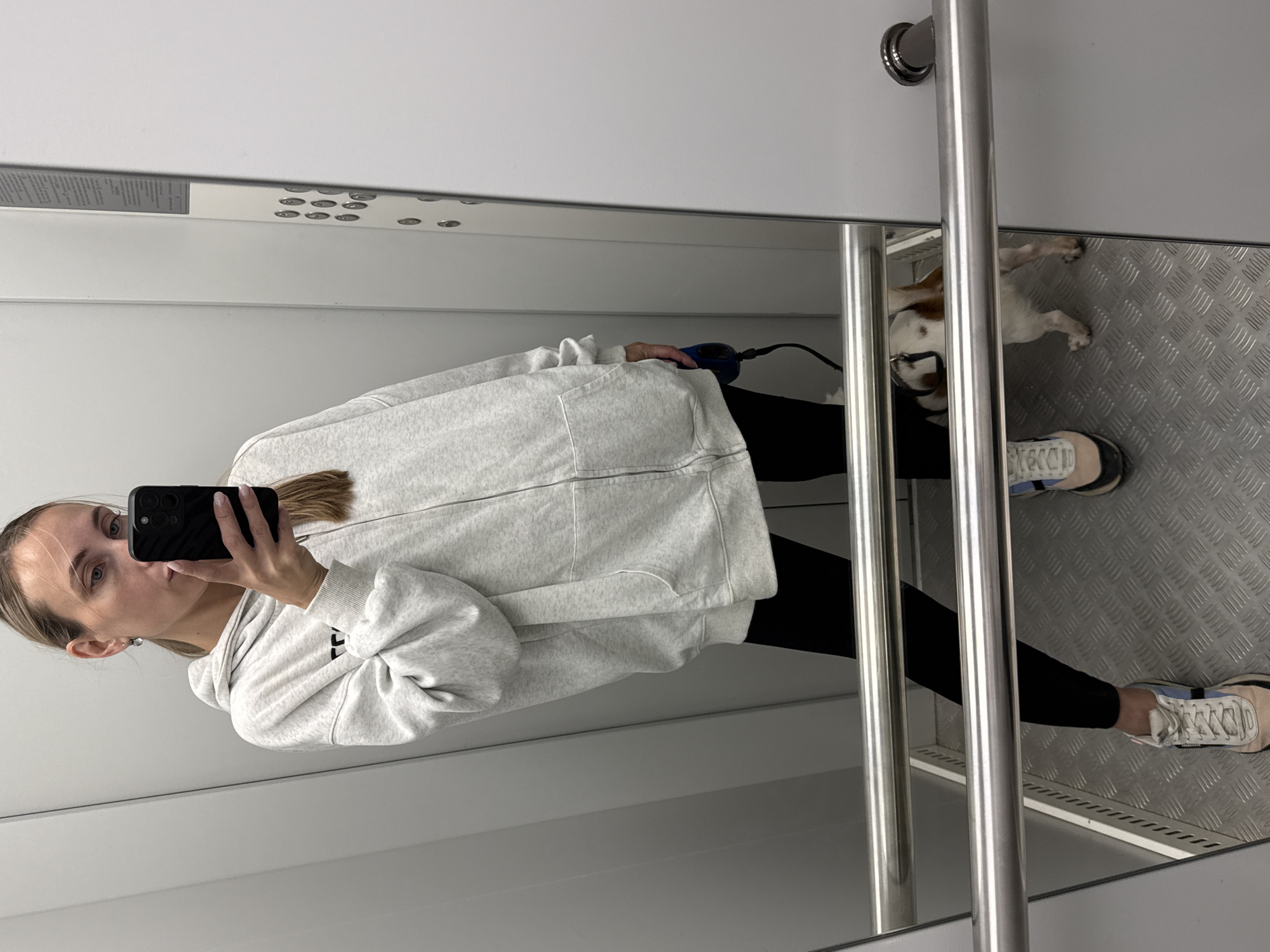A woman is taking a selfie in an elevator. She is wearing a light gray zip-up hoodie over black leggings and colorful sneakers. Her blonde hair is pulled back in a ponytail. She is holding a black smartphone in her right hand. A small dog with white and brown markings is visible in the reflection of the elevator door, standing on the diamond-plate floor. The elevator has stainless steel railings and a panel with buttons on the right wall. The image appears to have been taken indoors, likely in a residential building or a public facility in Chaikovsky, Russia.