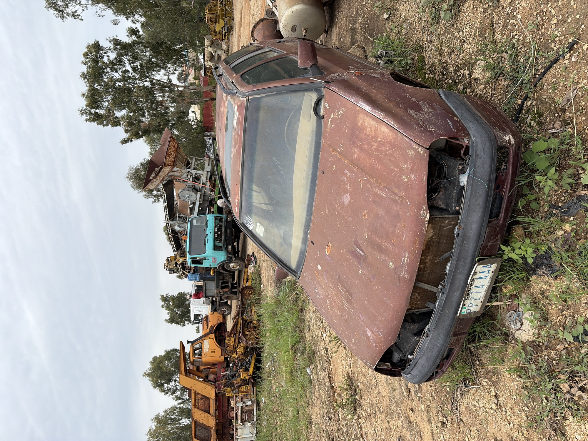 The image depicts a derelict, maroon-colored car in a junkyard or scrapyard setting in Gura Top, Nigeria. The car is heavily rusted and missing parts, including its headlights. The license plate is partially visible, bearing the inscription "PP274AAA" and "PLATEAU STATE-PEACE HOME OF TOURISM." The car is surrounded by dirt, vegetation, and various pieces of scrap metal and machinery. In the background, there are other vehicles, including trucks, and various pieces of industrial equipment. The sky is overcast.