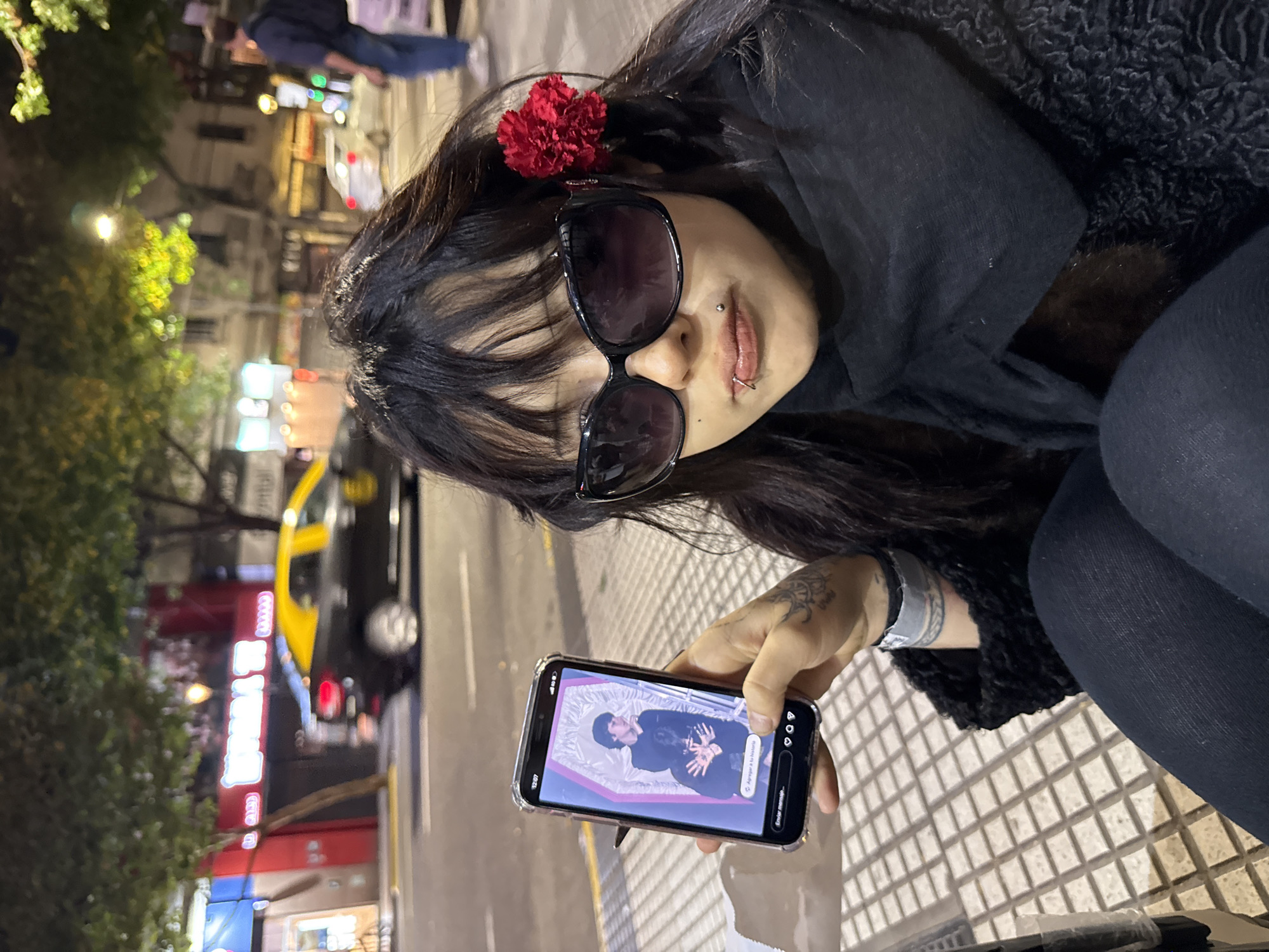 A young woman is sitting on a sidewalk in Buenos Aires, Argentina. She is wearing sunglasses, a black scarf, a black top, and black leggings. She has a red flower in her hair, a lip piercing, and tattoos on her left arm. She is holding a smartphone displaying an image of a man lying in a coffin. A taxi is parked on the street behind her, and storefronts are visible in the background. The sidewalk has a geometric pattern of light-colored tiles. The street lights illuminate the scene at night.