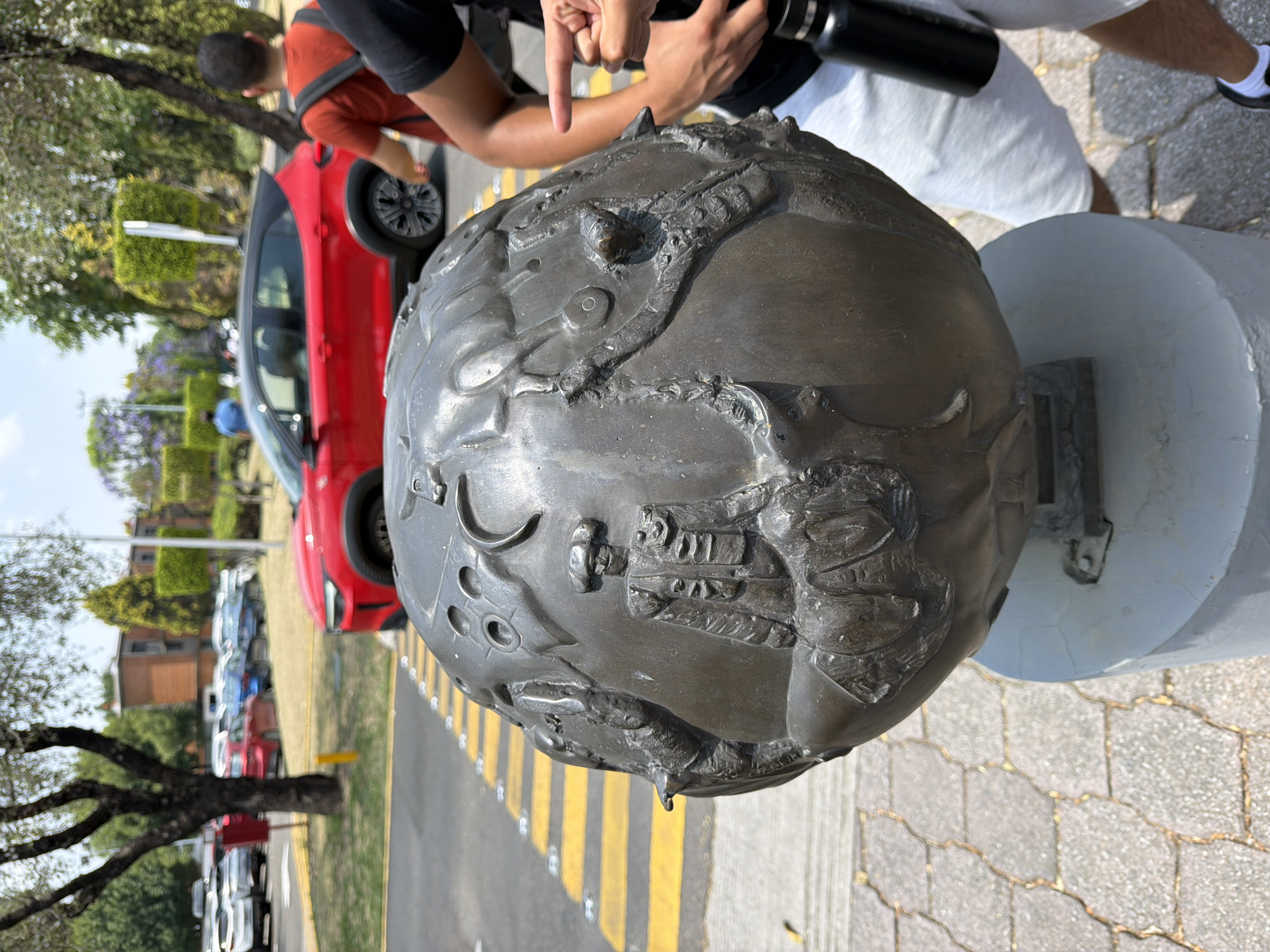 The image shows a bronze globe-shaped sculpture on a white pedestal in San Andrés Cholula, Mexico. The sculpture is detailed with raised designs and appears to be positioned on a brick walkway next to a street with yellow and black stripes. A person is pointing at the globe, while another person is holding a black water bottle. A red car and other parked vehicles are visible in the background. The background also includes trees, greenery, and buildings. The image is taken during daylight hours.
